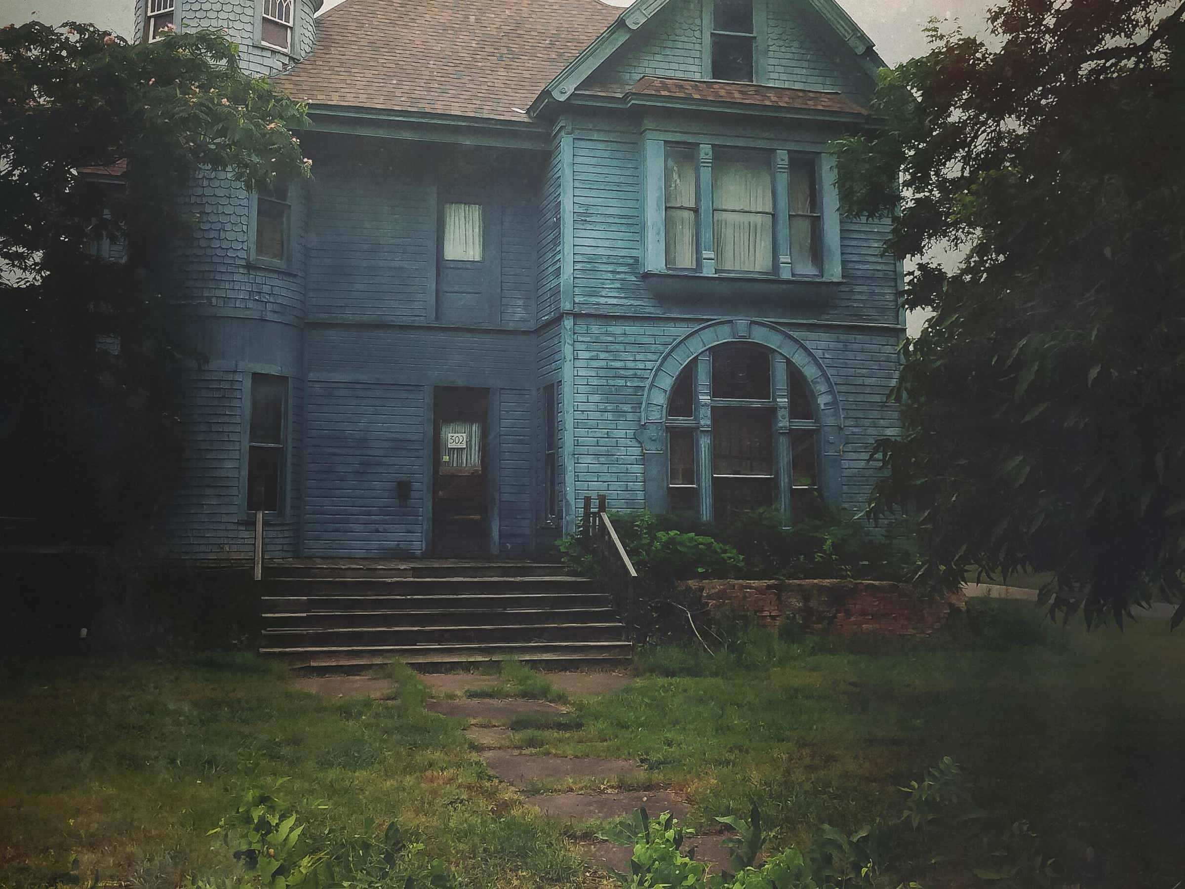 Haunted House 桌布 壁紙 — 一間風化的藍色維多利亞式房屋，油漆剝落，庭院雜草叢生，在陰沉天空下莊嚴地矗立。