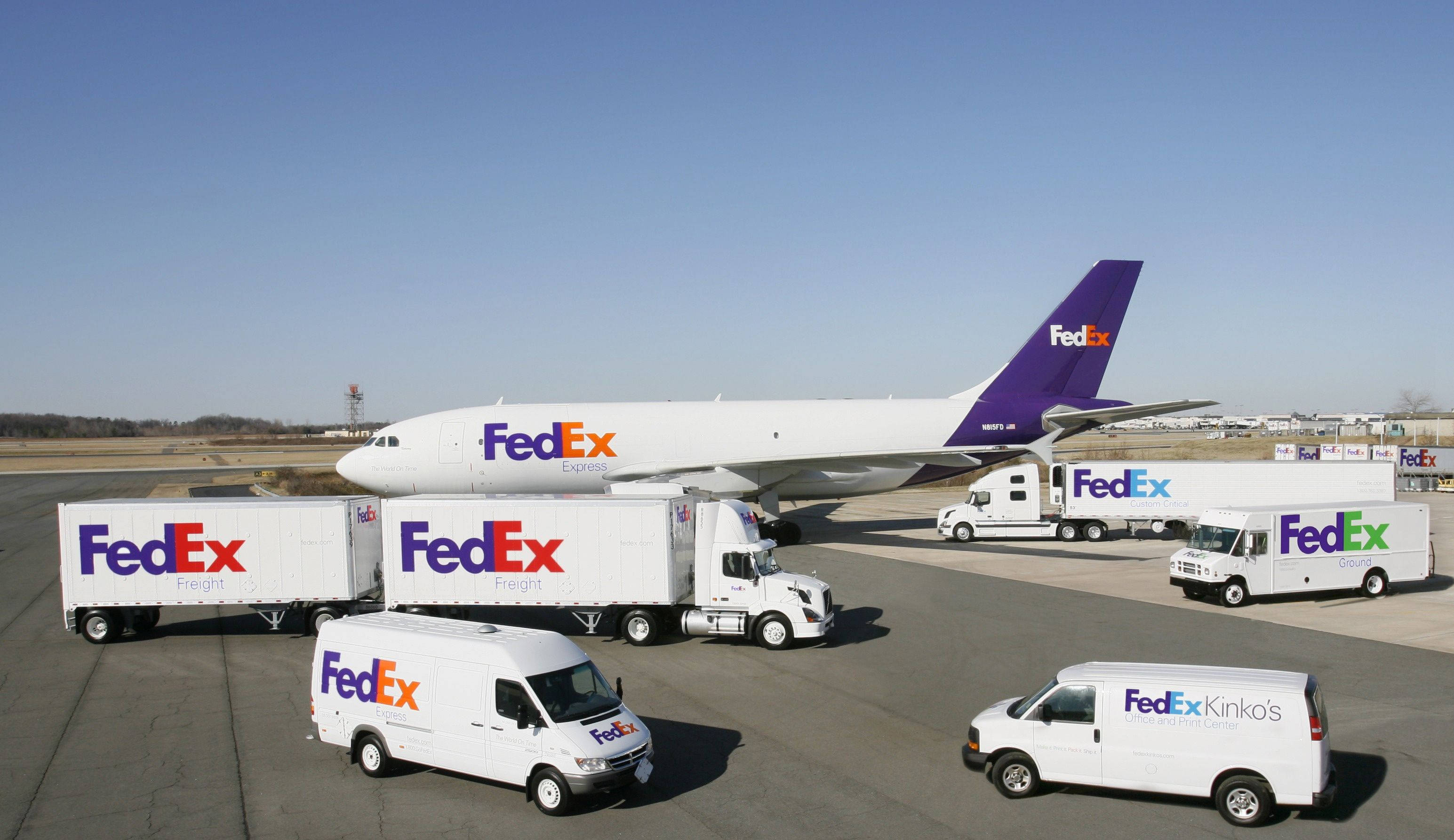 聯邦快遞 桌布 壁紙 — 一隊FedEx貨車和一架大型FedEx貨機停泊在機場停機坪上，天空湛藍。