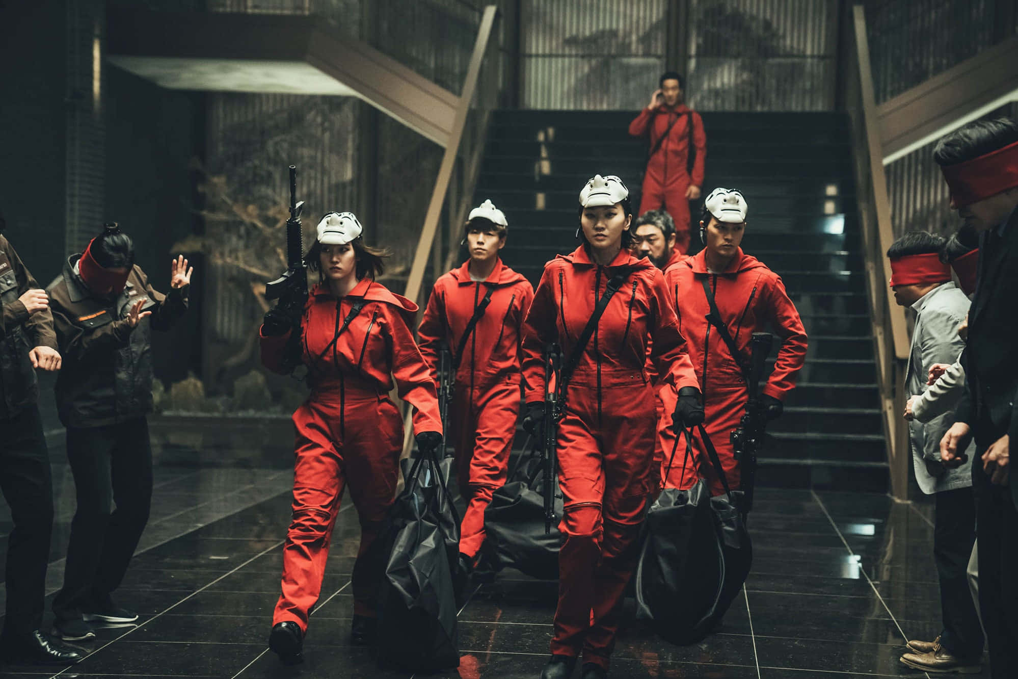 紙房子 桌布 壁紙 — 一隊身穿紅色連身衣褲和白色頭盔的女性，手持武器和袋子，沿著燈光昏暗的現代樓梯行進。