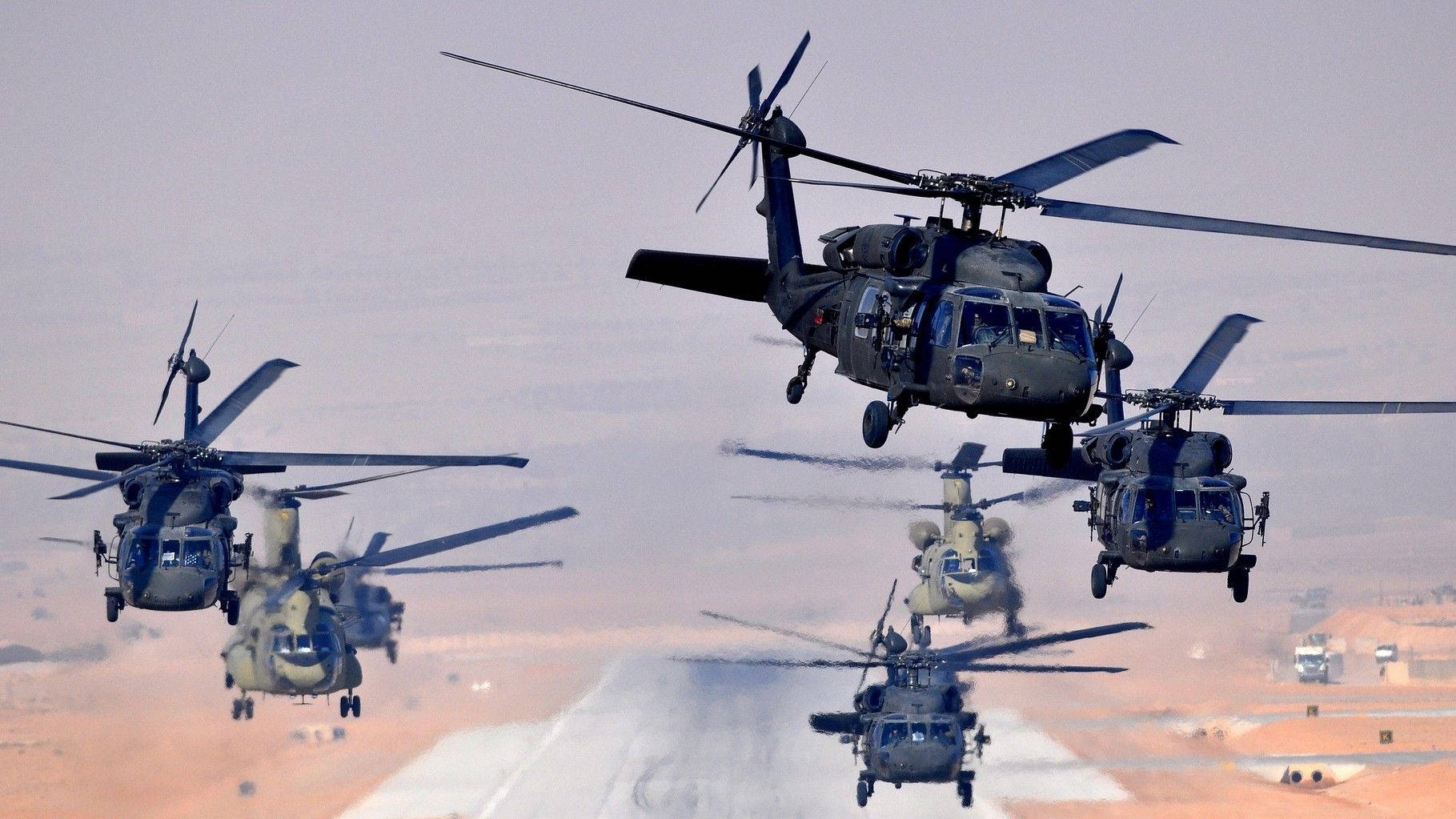 Black Hawk Helicopter 桌布 壁紙 — 一隊軍用直升機低飛越過沙漠跑道，在朦朧的天空中揚起塵土。
