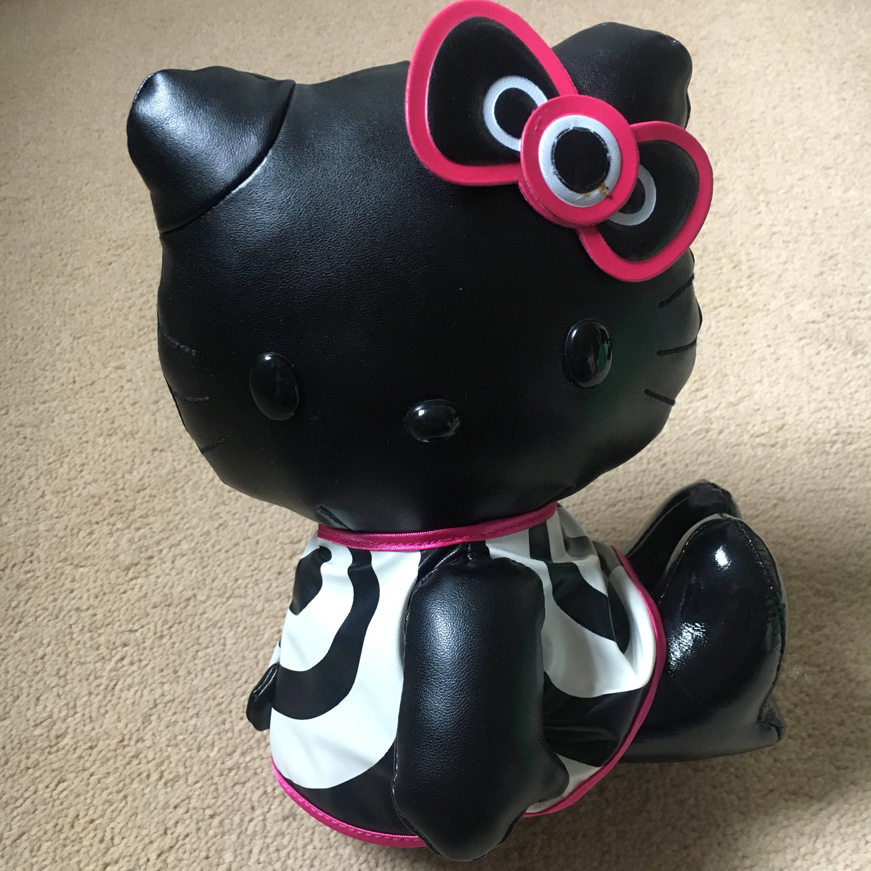 Hello Kitty Halloween 桌布 壁紙 — 一隻亮面黑色Hello Kitty絨毛玩偶，戴著亮粉紅色蝴蝶結，身穿黑白乳牛紋套裝，坐在米色地毯上。