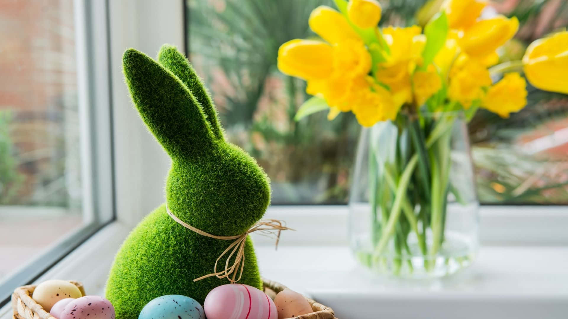 Easter Bunny 桌布 壁紙 — 一隻佈滿苔蘚的復活節兔子坐在陽光普照的窗台上，旁邊放著一籃色彩繽紛的復活蛋和一瓶鮮黃色的鬱金香。