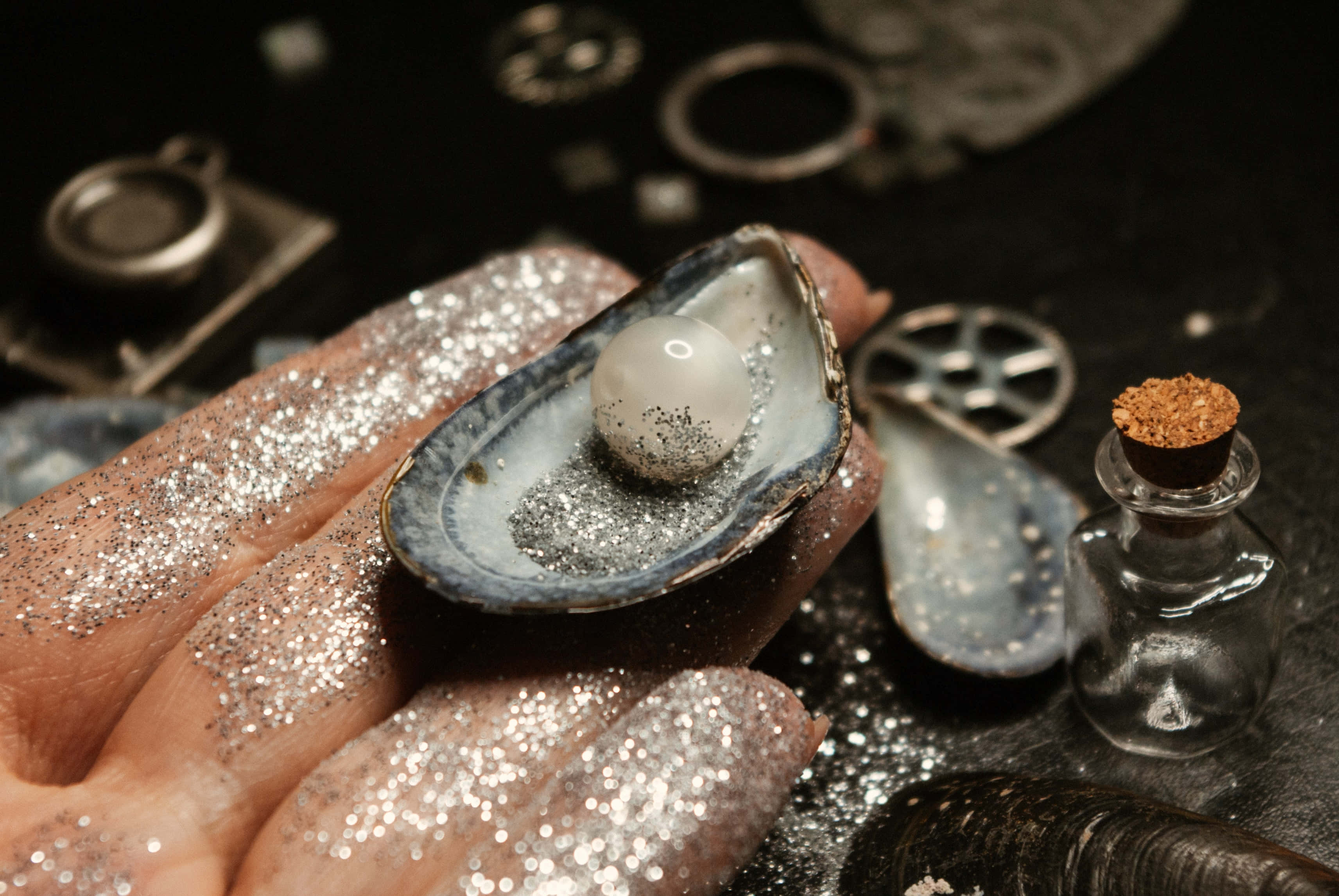 pearl 桌布 壁紙 — 一隻佈滿銀色亮粉的手，捧著一個打開的牡蠣殼，裡面有一顆發光的珍珠，背景是深色的，散落著貝殼和一個小小的軟木塞瓶子。