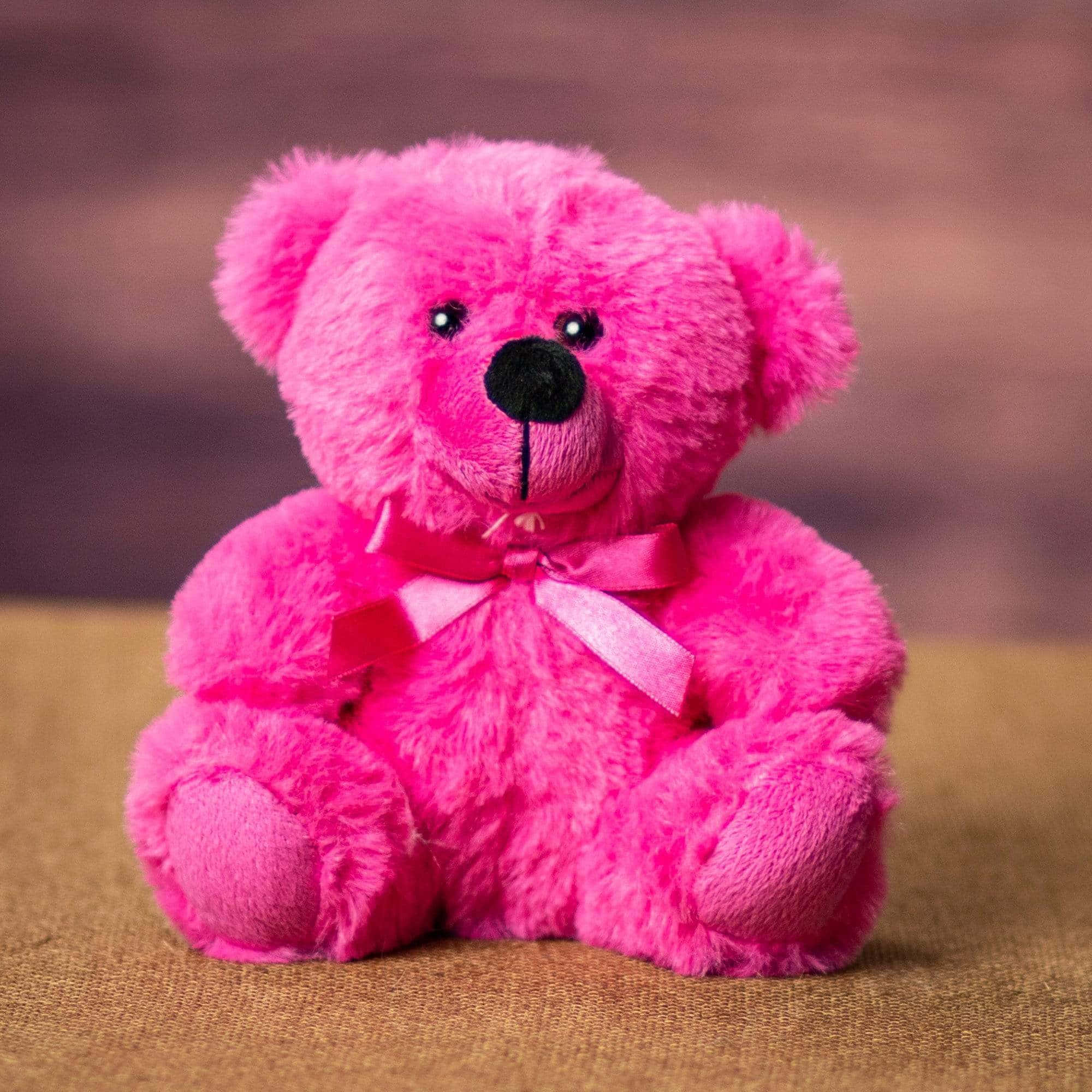 Teddy Bear 桌布 壁紙 — 一隻充滿活力的粉紅色毛絨泰迪熊，戴著相配的蝴蝶結，坐在有紋理的棕色表面上，背景是模糊的暖色調。