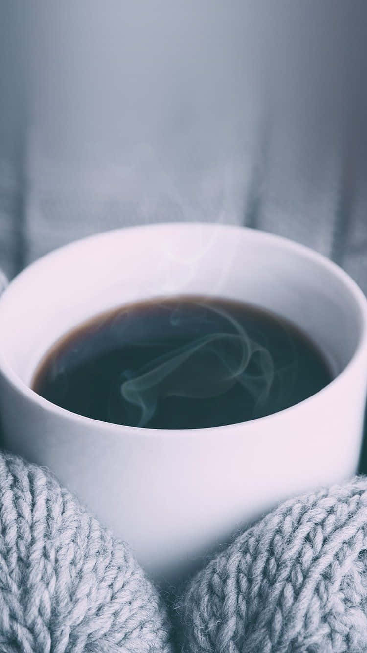 冬季美學 桌布 壁紙 — 一隻冒著蒸氣的白色馬克杯裝著黑咖啡，被柔軟的灰色針織手套包裹，背景是柔和的灰色。