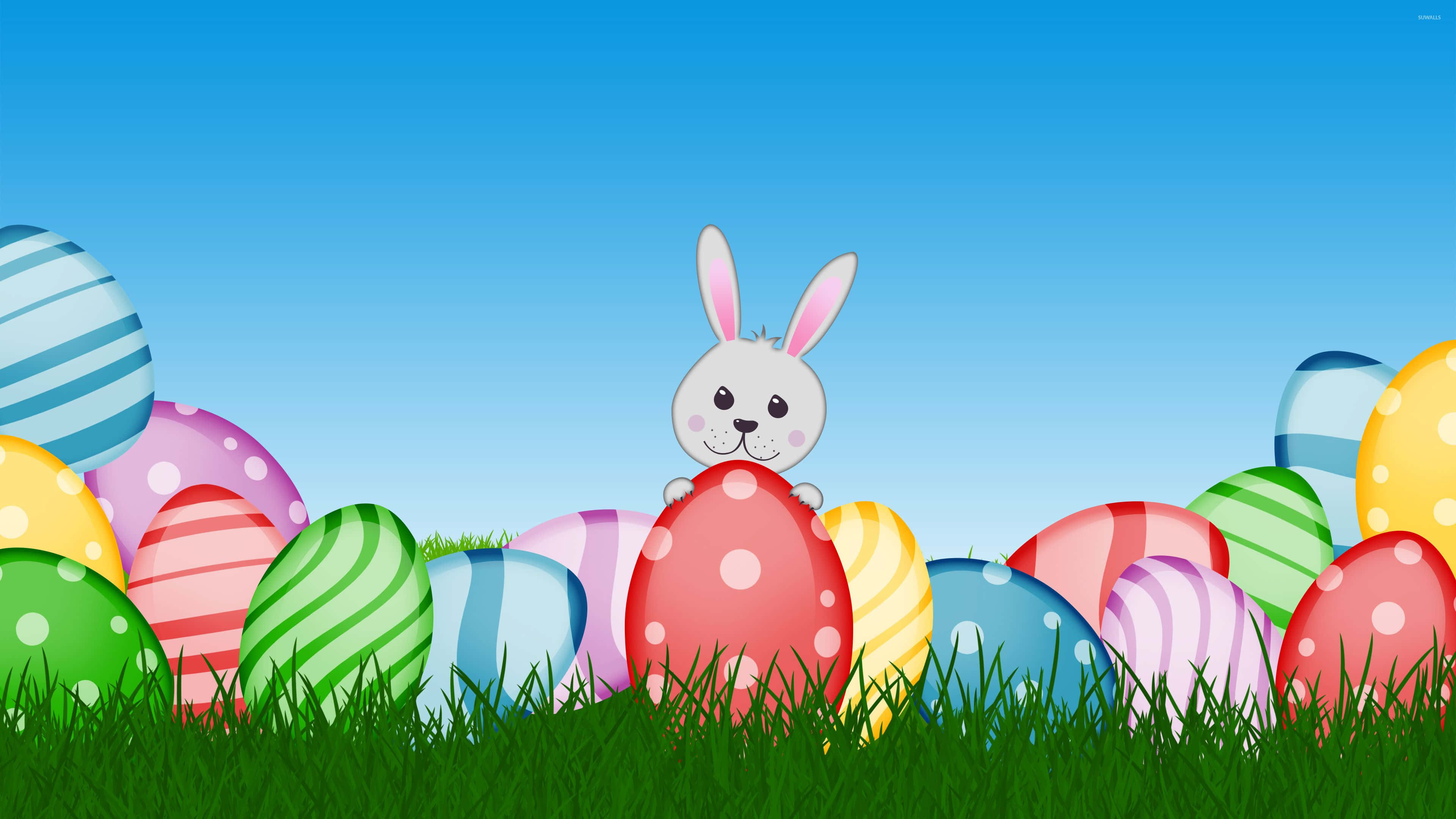 Easter Bunny 桌布 壁紙 — 一隻可愛的白兔從色彩繽紛的復活節蛋上方探出頭來，這些蛋安放在晴朗藍天下茂盛的綠草中。