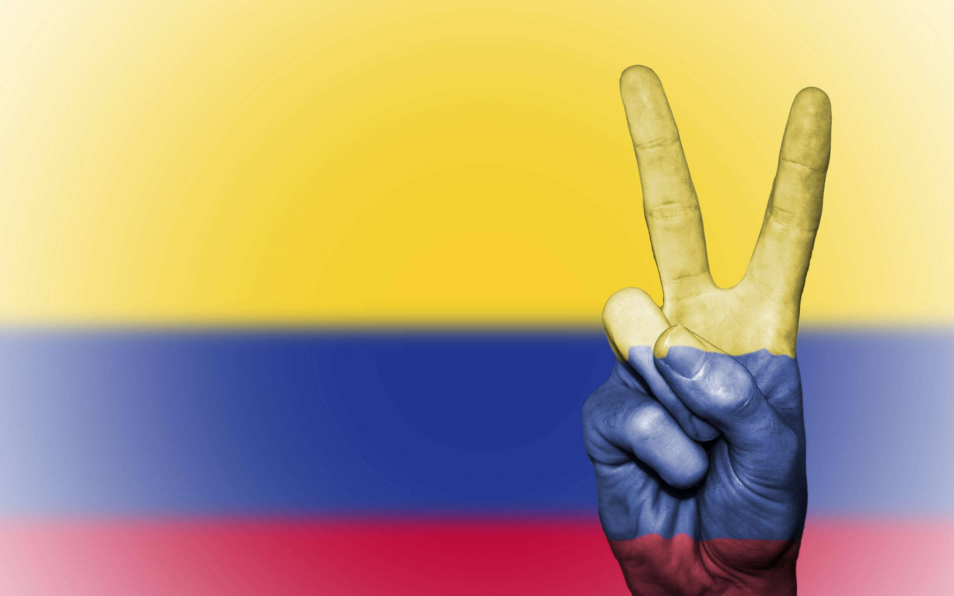 Colombia Flag 桌布 壁紙 — 一隻塗有哥倫比亞國旗顏色的手，在模糊的國旗背景下做出和平手勢。