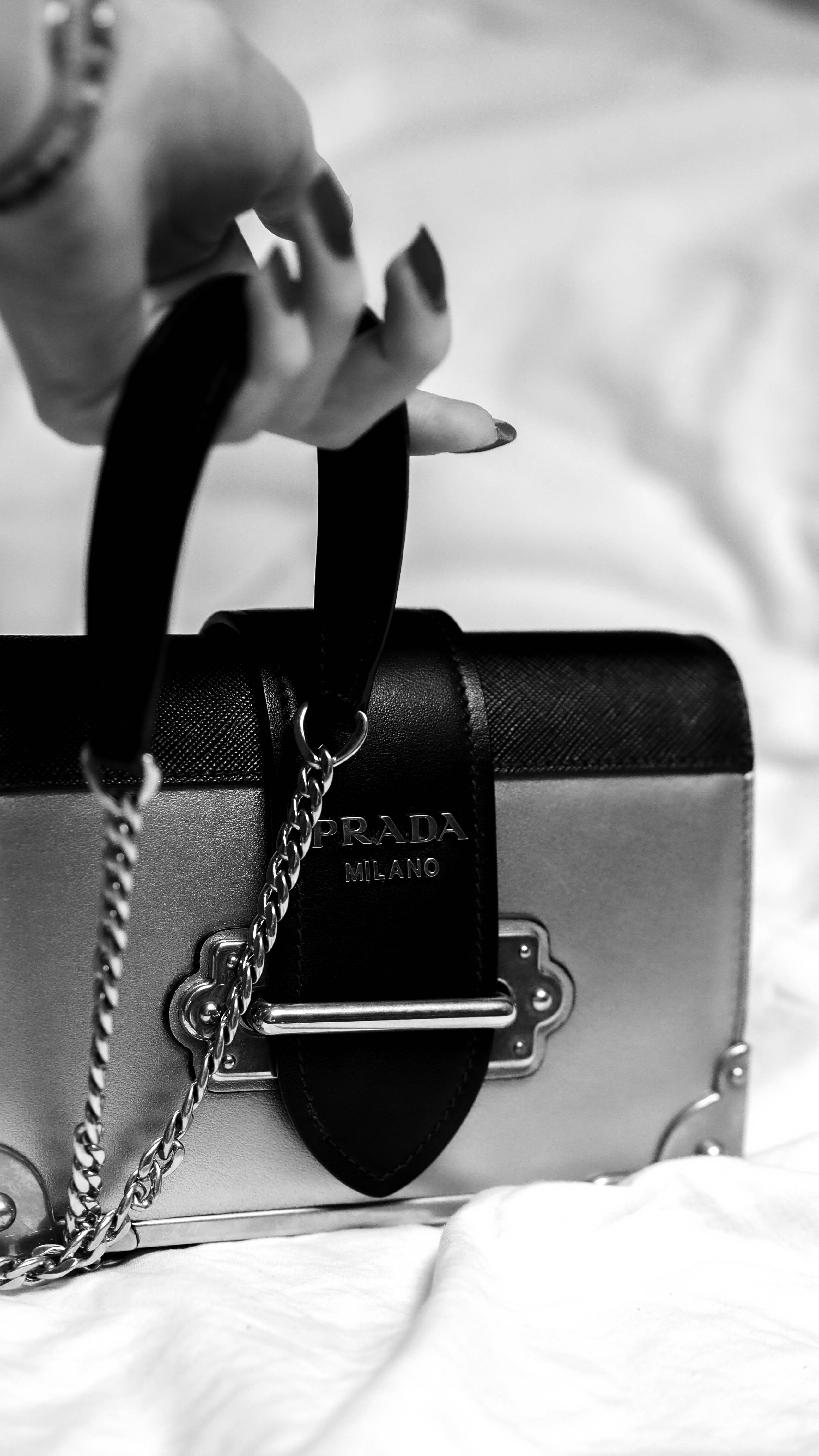 Prada 桌布 壁紙 — 一隻塗有深色指甲油的手，在柔軟的白色布料背景上，以單色調拿著一個黑色和銀色的Prada手袋。