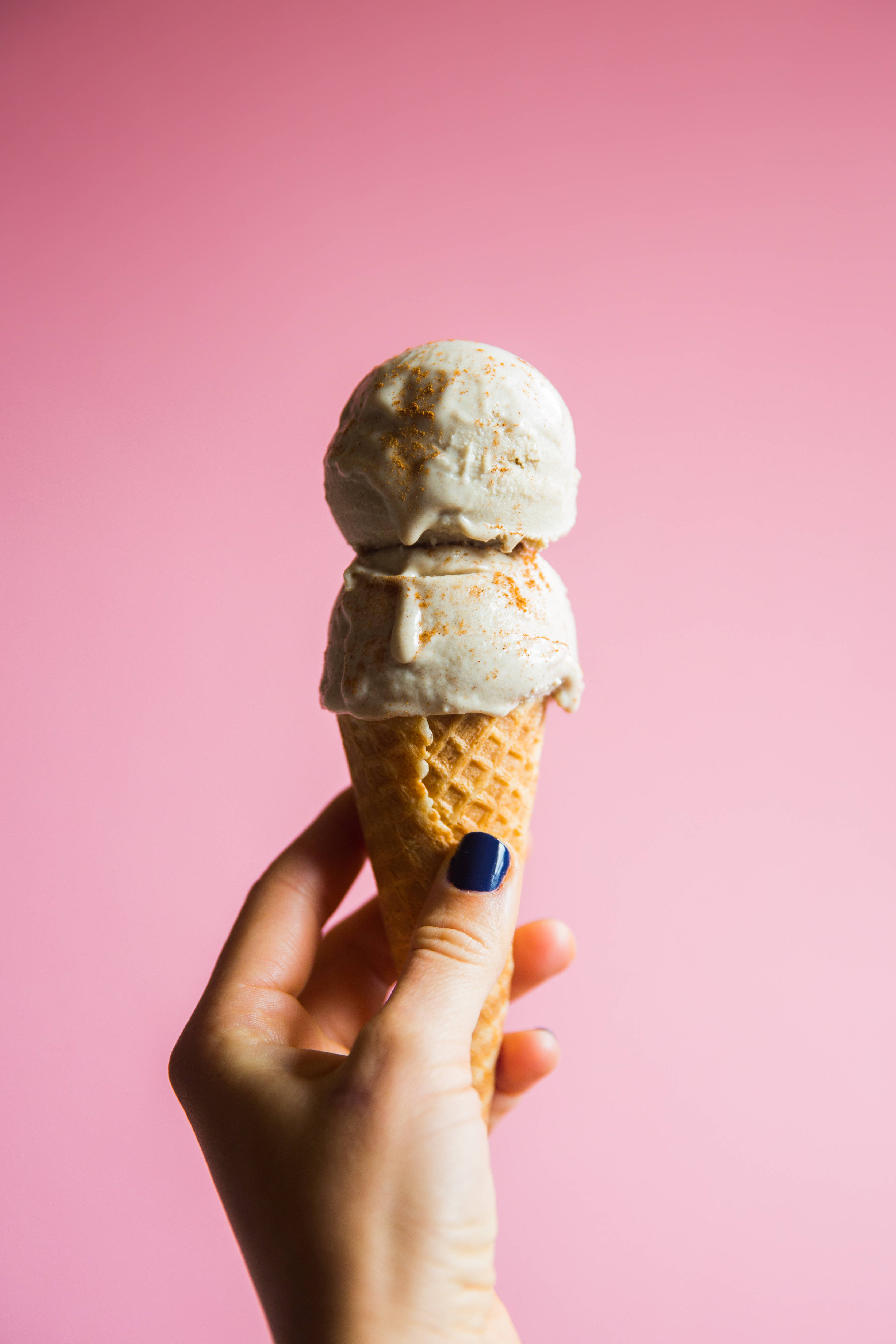 Ice Cream 桌布 壁紙 — 一隻塗有深藍色指甲油的手拿著一個鬆餅筒，上面有兩球肉桂冰淇淋，背景是柔和的粉紅色。