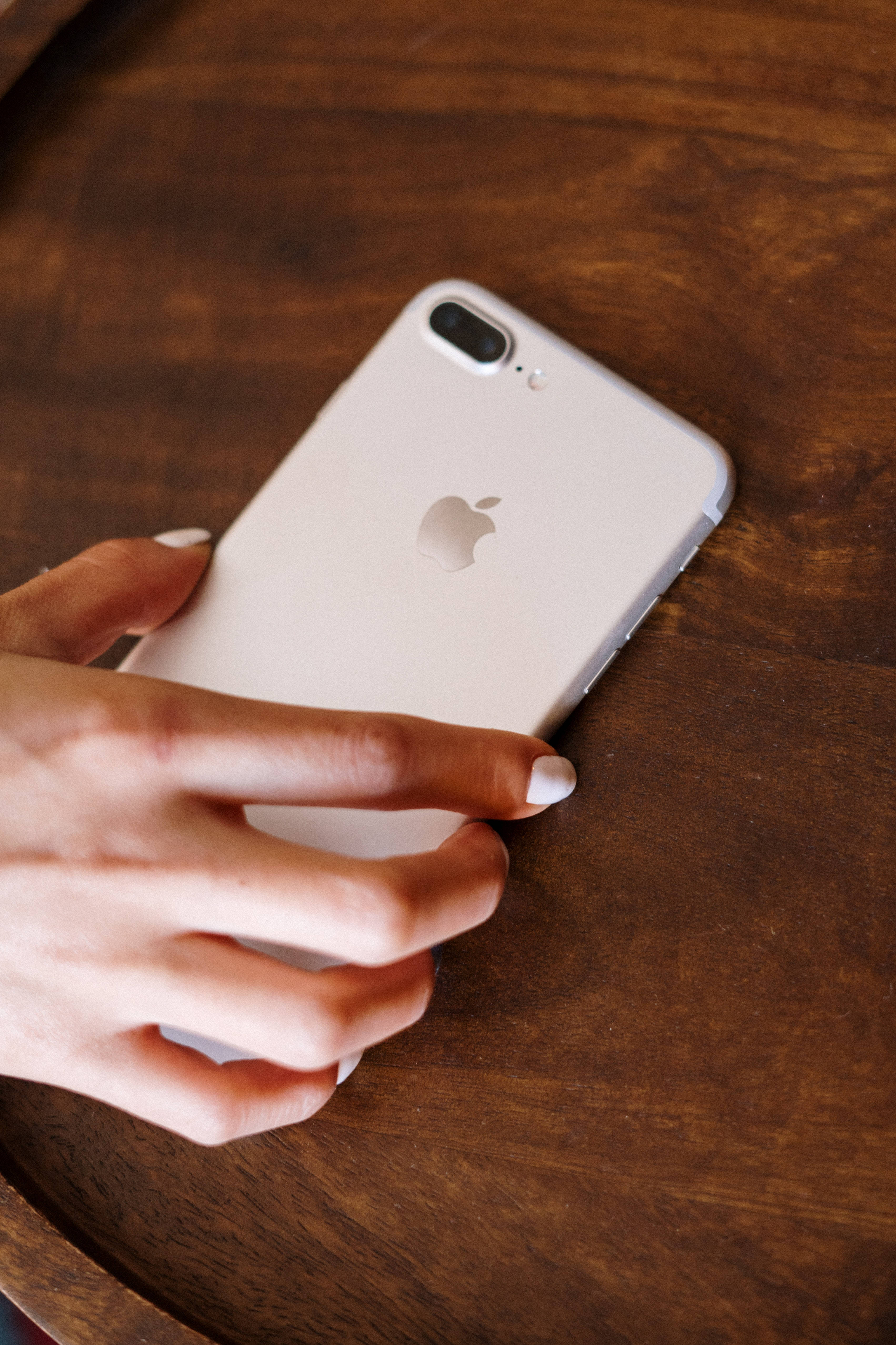 iPhone 桌布 壁紙 — 一隻塗有白色指甲油的手輕輕地拿著一部銀色iPhone放在一張豐富的棕色木桌上。