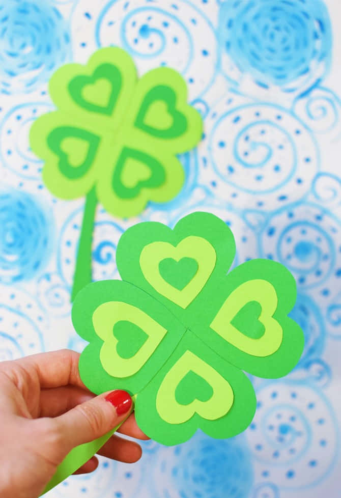 Four Leaf Clover 桌布 壁紙 — 一隻塗有紅色指甲油的手，拿著兩片帶有心形鏤空的亮綠色紙質三葉草，背景是藍色漩渦圖案。
