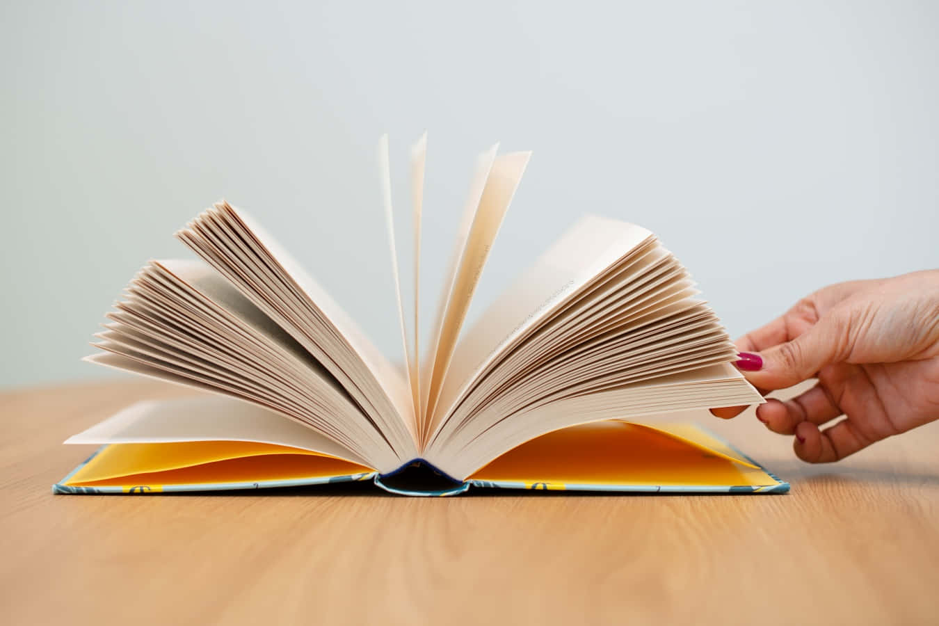 Open Book 桌布 壁紙 — 一隻塗有紅色指甲油的手輕輕翻動放在淺色木桌上的一本打開的書的書頁。