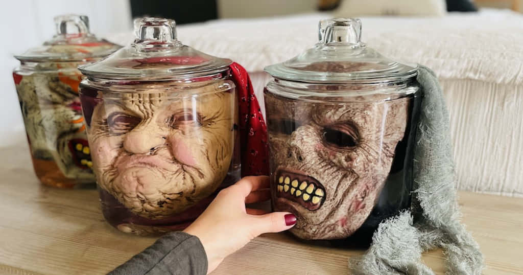 Creepy Halloween 桌布 壁紙 — 一隻塗有紅色指甲的手輕輕觸摸一個裝有保存殭屍面具的玻璃罐，旁邊的木桌上還有另外兩個罐子。