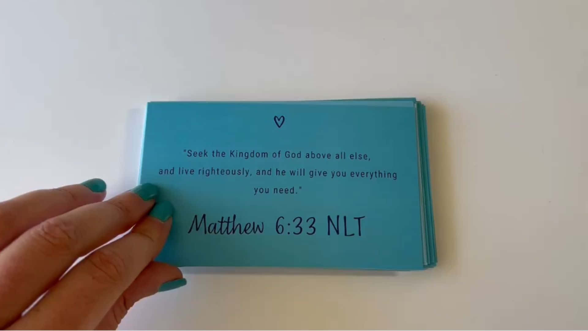Scripture 桌布 壁紙 — 一隻塗有青綠色指甲油的手，在純白色背景前拿著一疊淺藍色卡片，卡片上印有馬太福音 6:33 NLT 的經文。