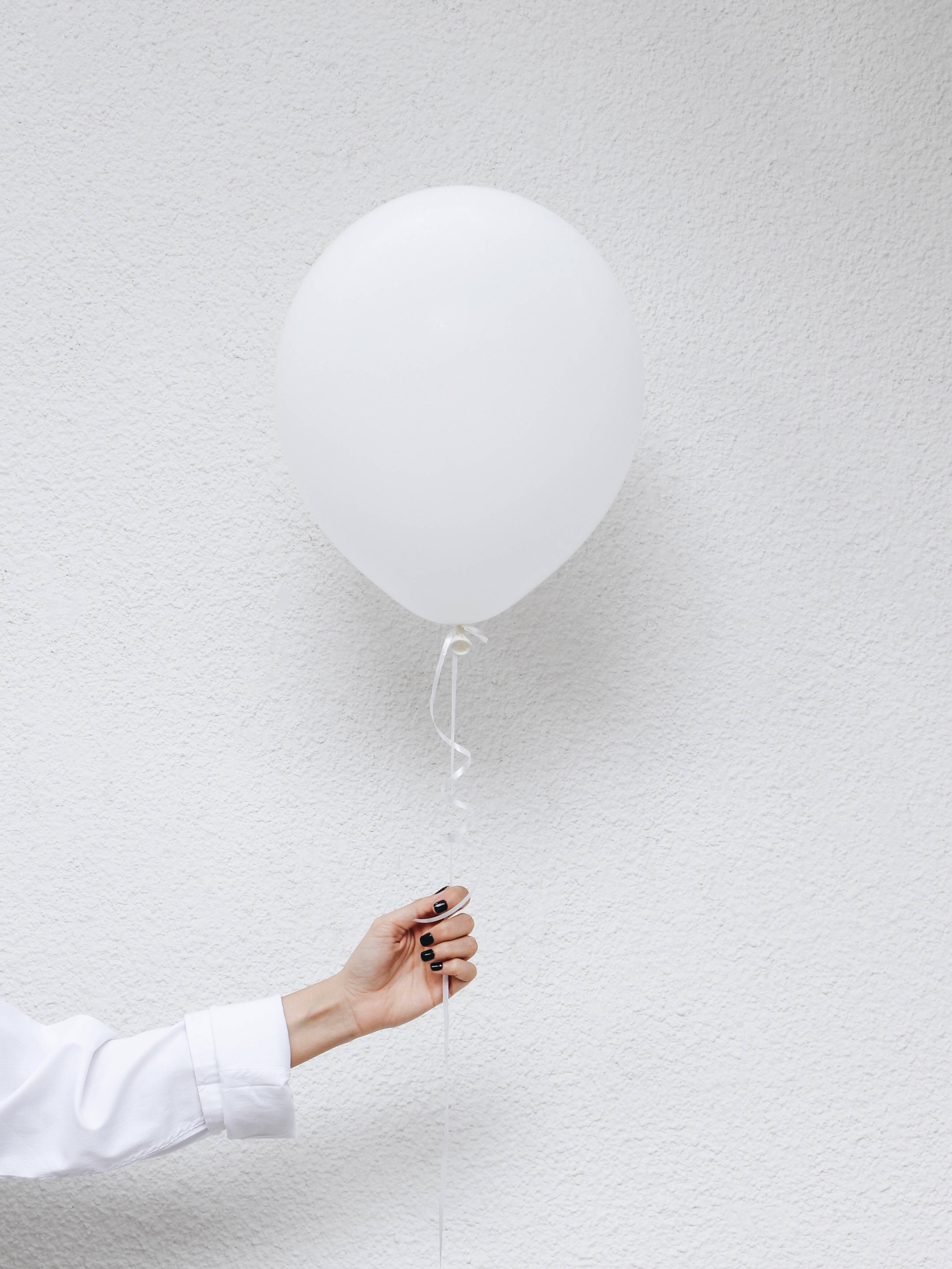 Balloon 桌布 壁紙 — 一隻塗有黑色指甲油的手拿著一個白色氣球，靠著一面有紋理的白色牆壁。