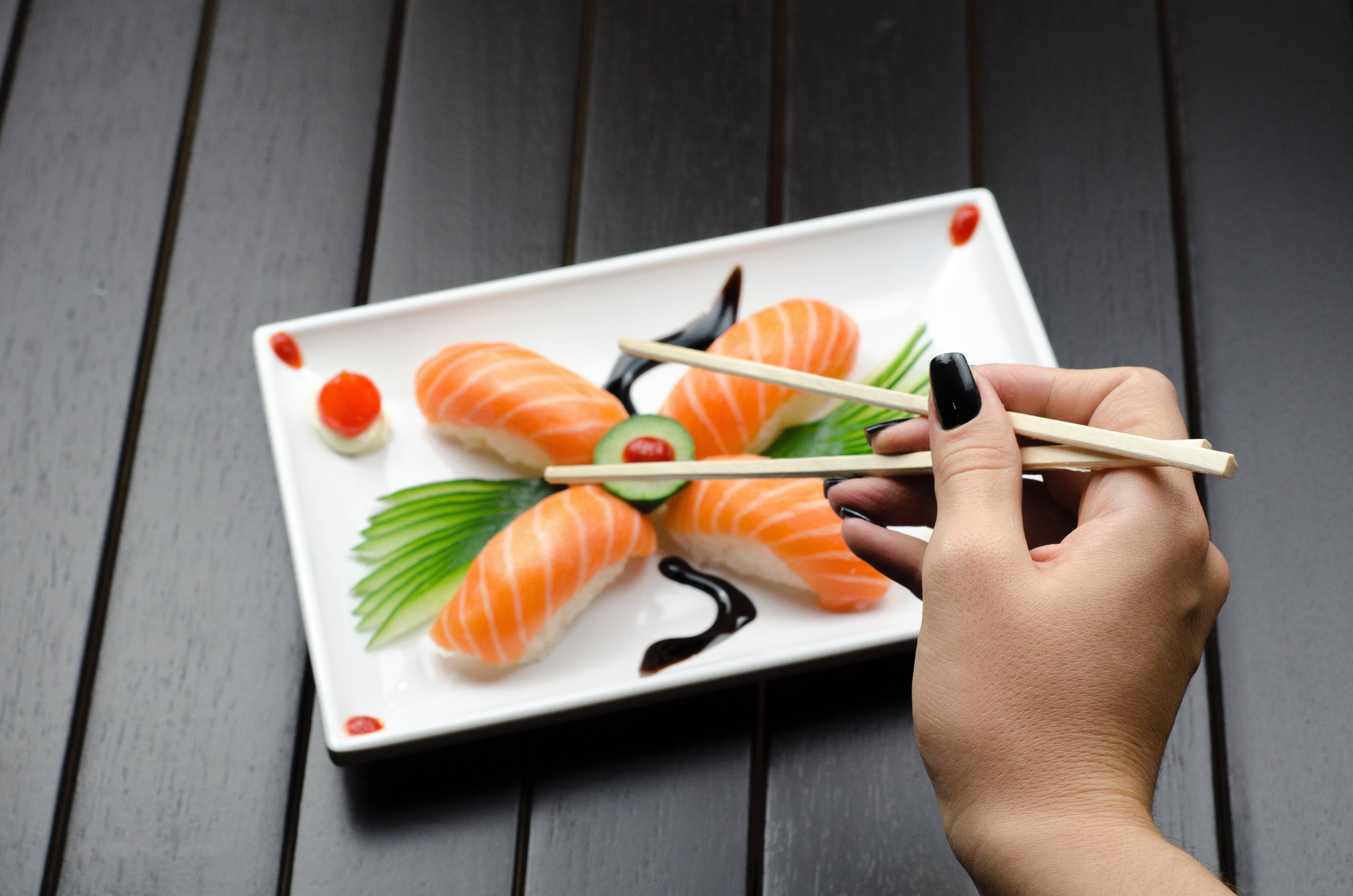 Sushi 桌布 壁紙 — 一隻塗有黑色指甲油的手用筷子從一個飾有芥末和醃薑的白盤中夾起鮭魚握壽司。