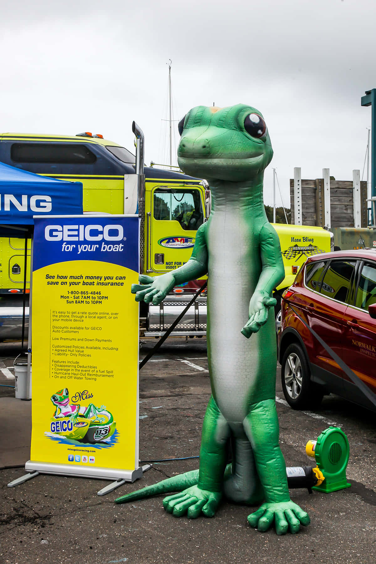 Geico 桌布 壁紙 — 一隻大型綠色壁虎吉祥物站在Geico船保險標誌旁邊，背景是戶外活動中的車輛和帳篷。