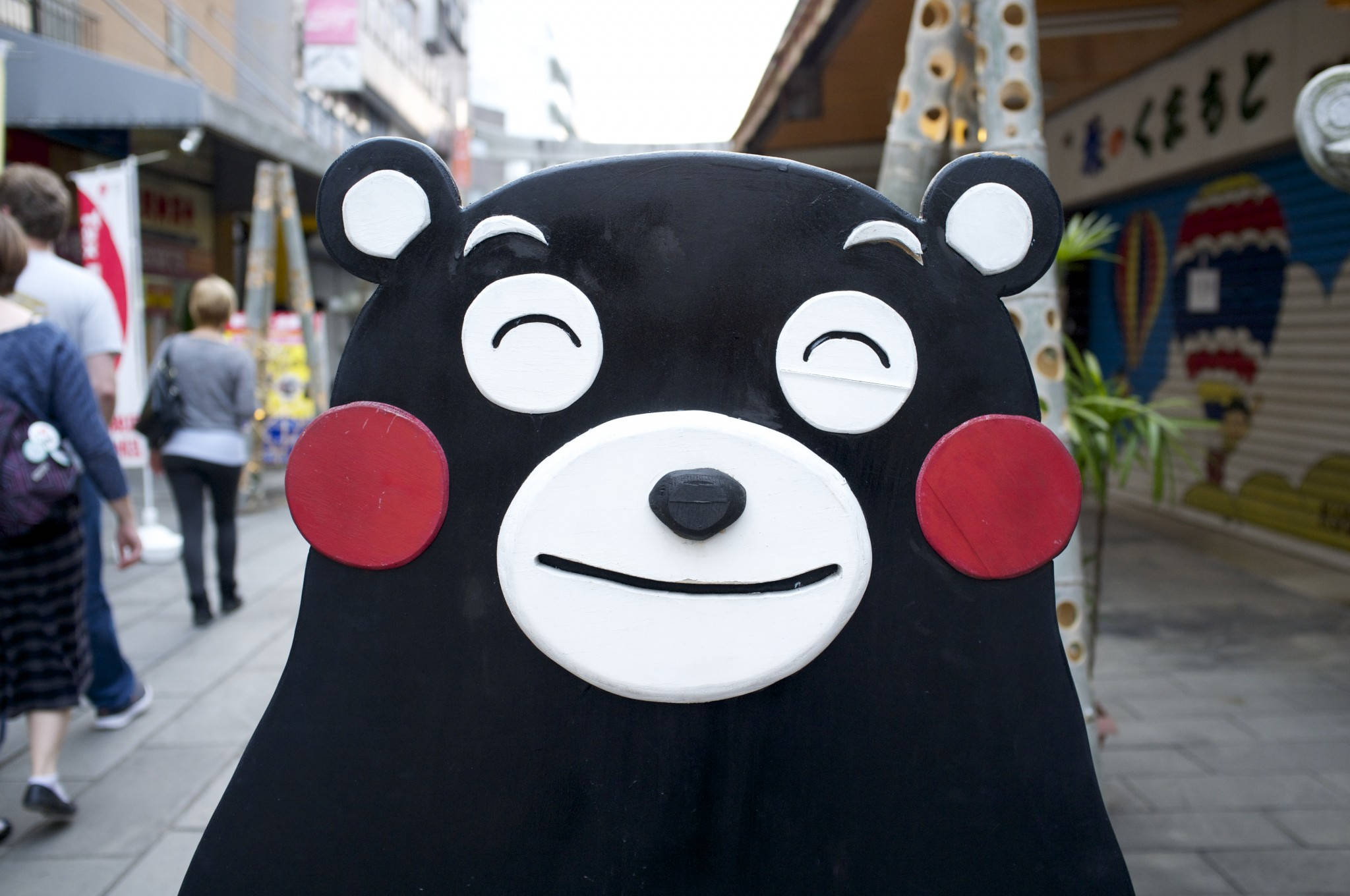 熊本熊 桌布 壁紙 — 一隻大型黑色熊吉祥物，臉頰紅潤、雙眼緊閉，站在繁忙的城市人行道上，行人來來往往。
