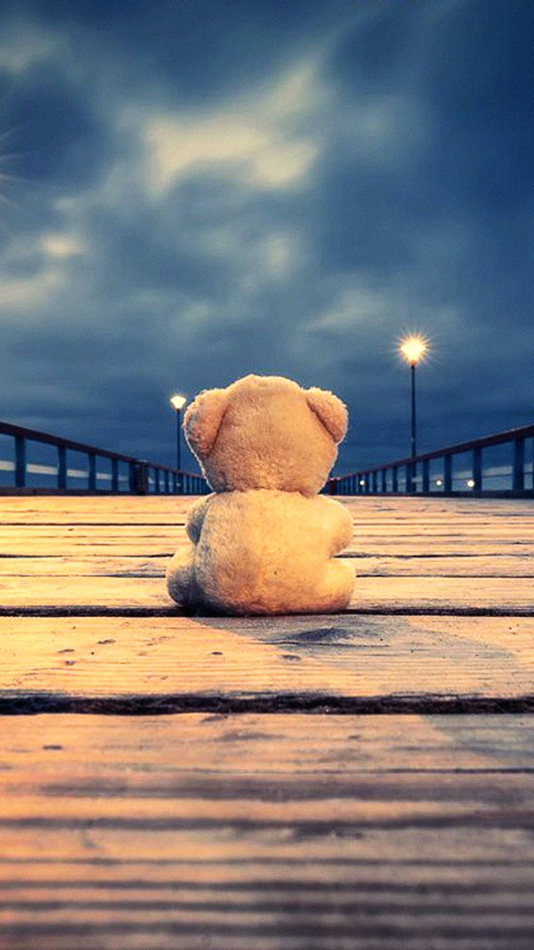 Teddy Bear 桌布 壁紙 — 一隻孤獨的米色泰迪熊坐在黃昏的木碼頭上，凝視著陰沉多雲天空下閃爍的街燈。