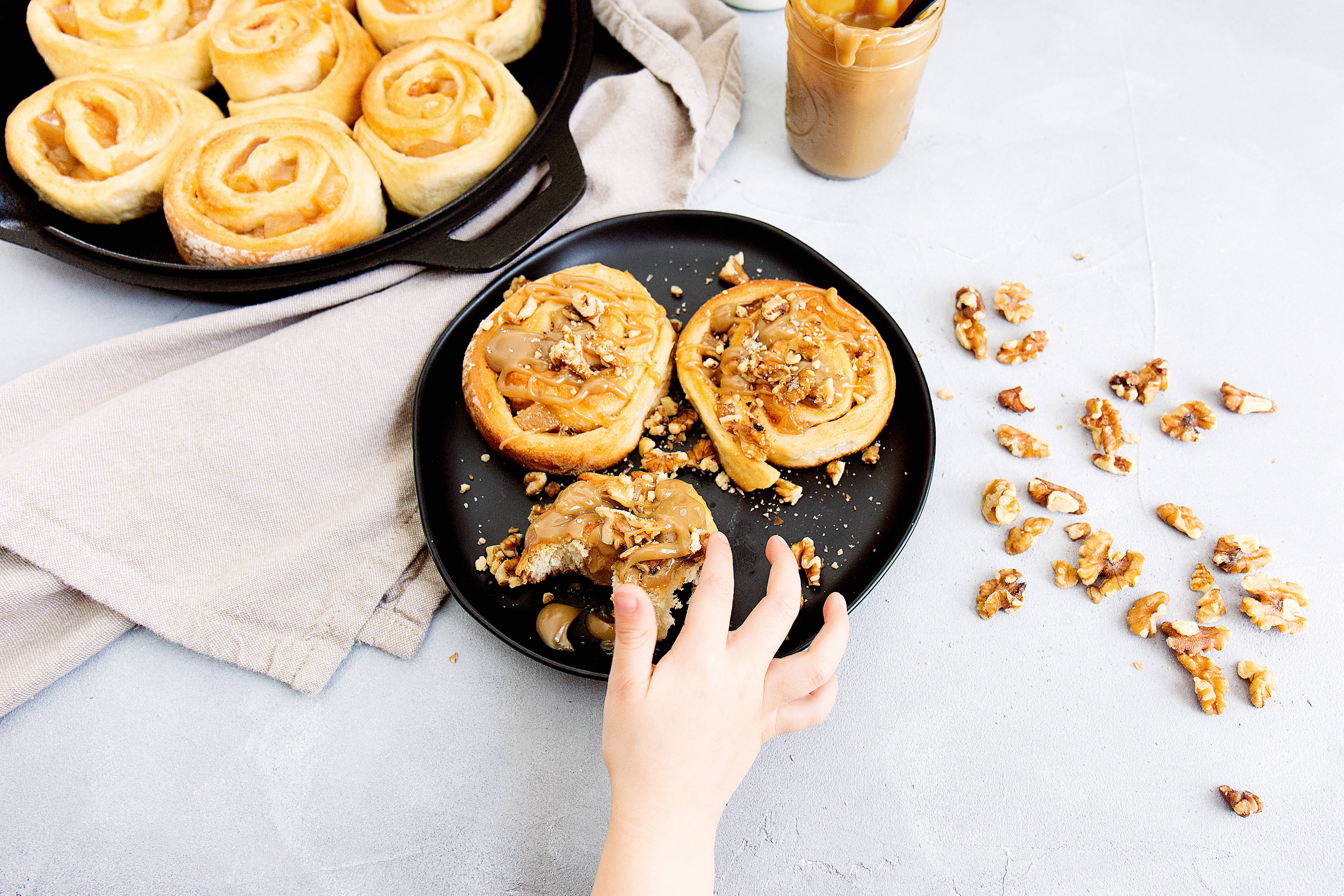 Pastry 桌布 壁紙 — 一隻孩子的手伸向黑色盤子上的焦糖核桃肉桂捲，旁邊的鑄鐵煎鍋裡還有更多肉桂捲。