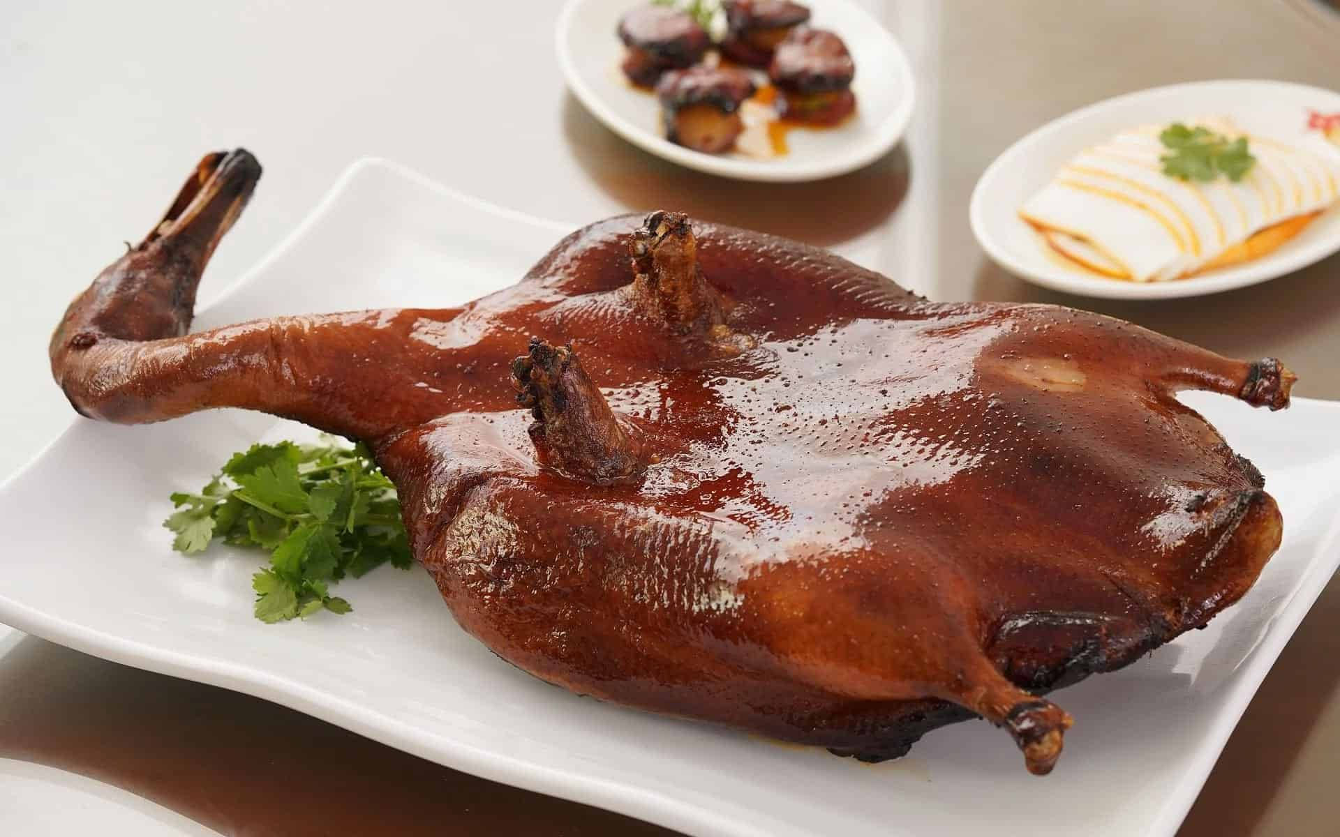 鴨子 桌布 壁紙 — 一隻完整光亮的烤鴨，皮脆肉嫩，放在白色盤子上，飾以新鮮香菜。