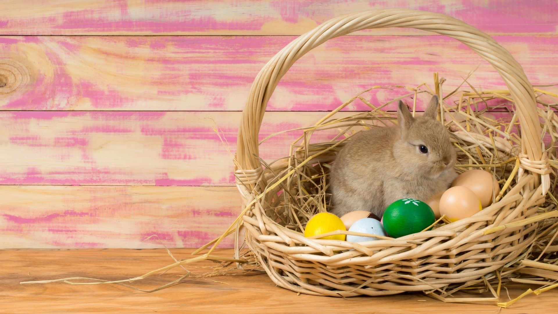 Easter Bunny 桌布 壁紙 — 一隻小棕色兔子坐在一個裝滿乾草和彩色復活節蛋的柳條籃子裡，背景是粉紅色的木質背景。