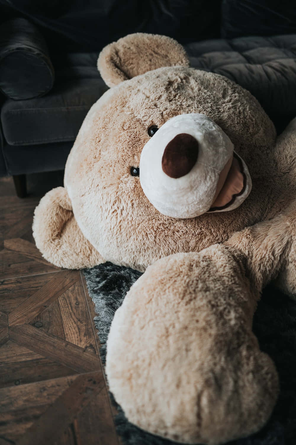 Teddy Bear 桌布 壁紙 — 一隻巨大毛絨米色泰迪熊舒適地躺在溫暖光線房間內深色地毯上，旁邊是皮革沙發。