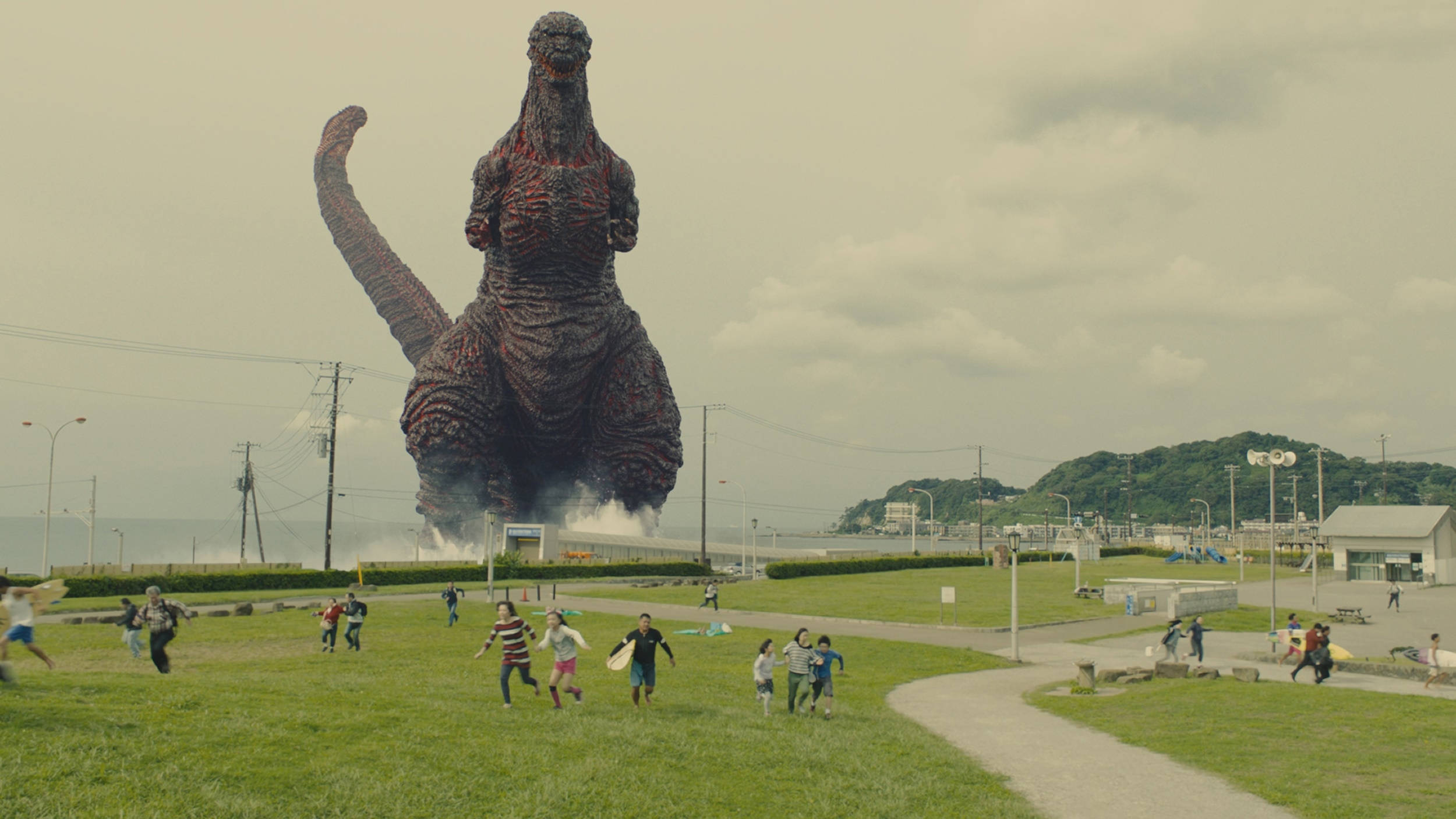 shin godzilla 桌布 壁紙 — 一隻巨大的哥吉拉從海中出現在一個草地公園，人們在陰天天空下驚恐逃離。