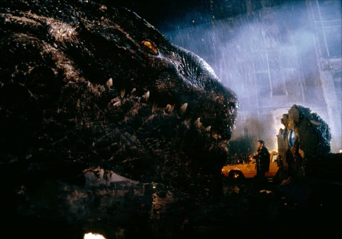 godzilla 1998 桌布 壁紙 — 一隻巨大的哥吉拉籠罩在被雨水浸透的城市街道上，一個孤獨的身影在黑暗中站在一輛黃色汽車旁邊。