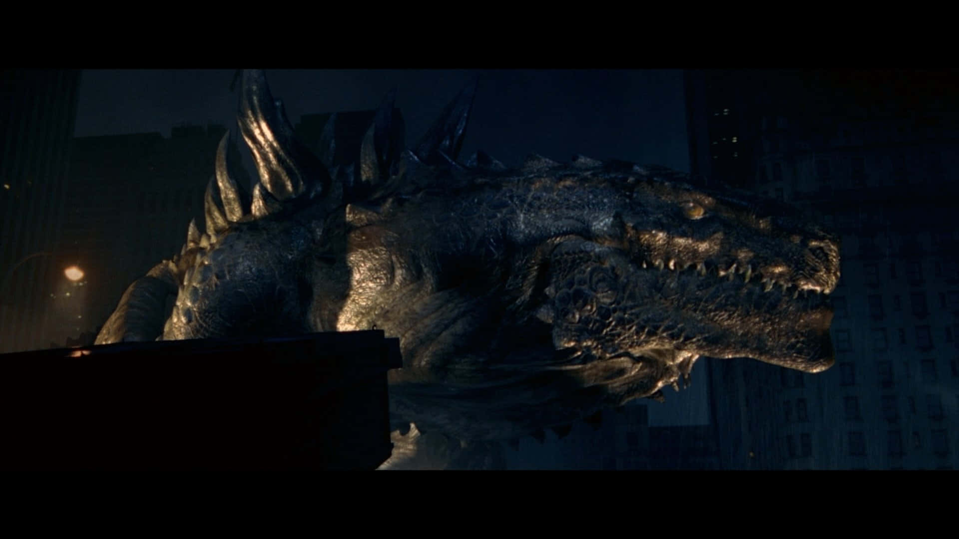 godzilla 1998 桌布 壁紙 — 一隻巨大的哥斯拉在夜晚籠罩著黑暗的城市景觀，在路燈下閃爍著發光的眼睛和鋒利的牙齒。