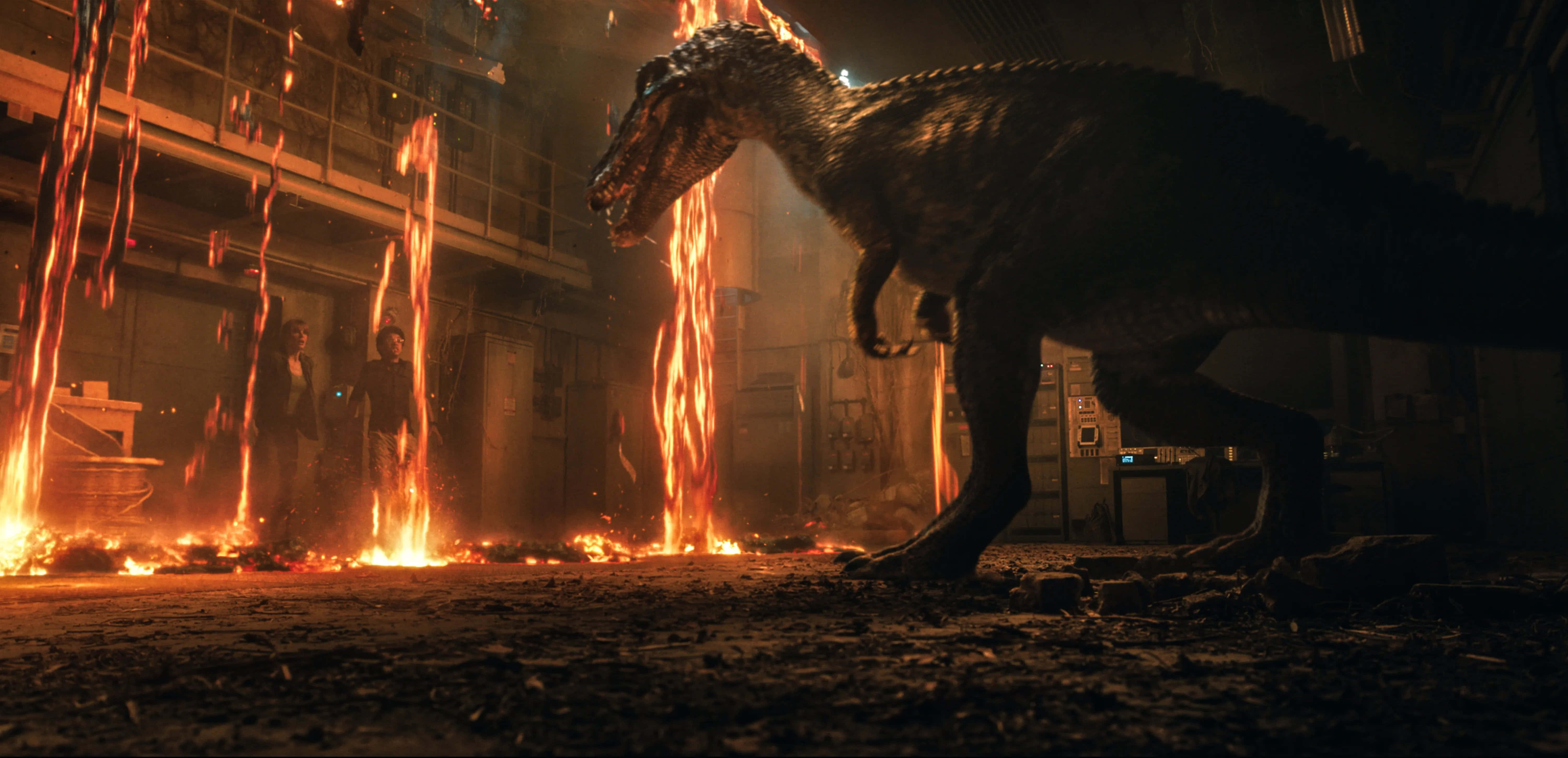 Jurassic World 桌布 壁紙 — 一隻巨大的恐龍站在黑暗工業廢墟中燃燒的瀑布之間，兩個人物在火焰後面觀看。