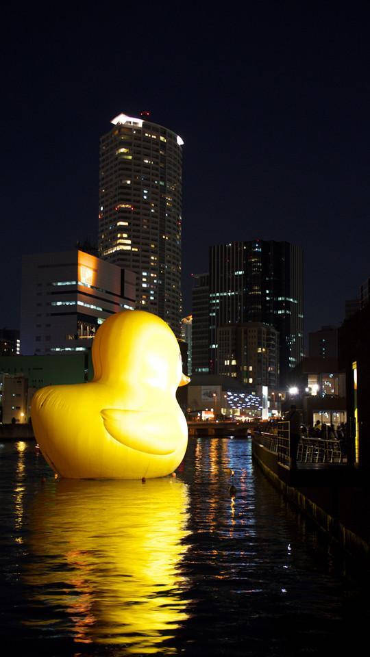 夜晚在城市建築附近的水面上漂浮的巨大發光黃色橡膠鴨。