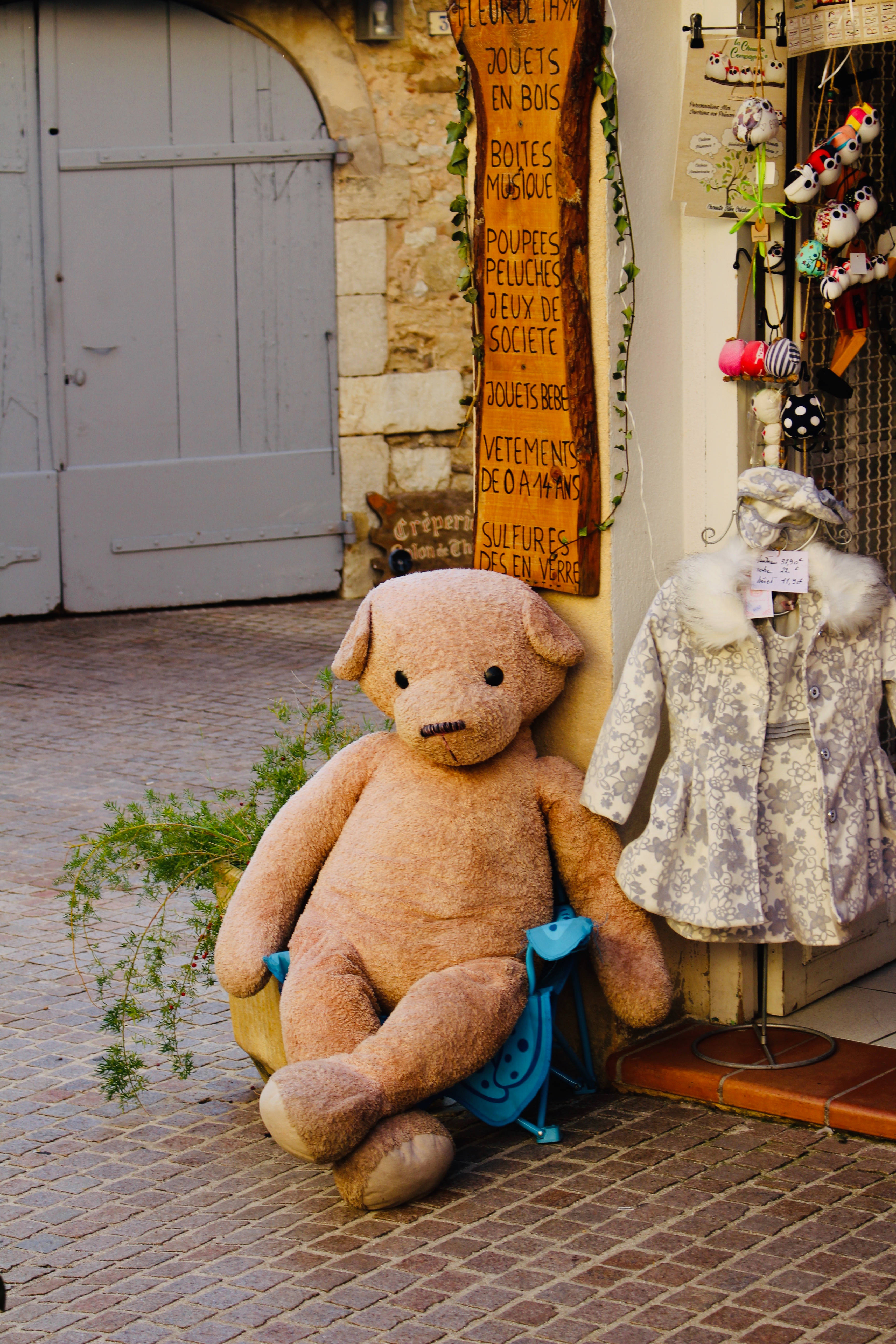 Cute Bear 桌布 壁紙 — 一隻巨大的米色泰迪熊坐在藍色凳子上，旁邊是兒童衣帽架，位於歐洲風格鵝卵石商店門口。