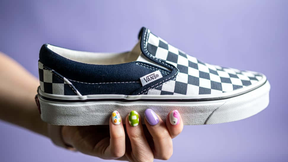 Vans 桌布 壁紙 — 一隻帶有彩色圓點指甲的手，在柔和的紫色背景前，拿著經典黑白格紋Vans slip-on運動鞋。