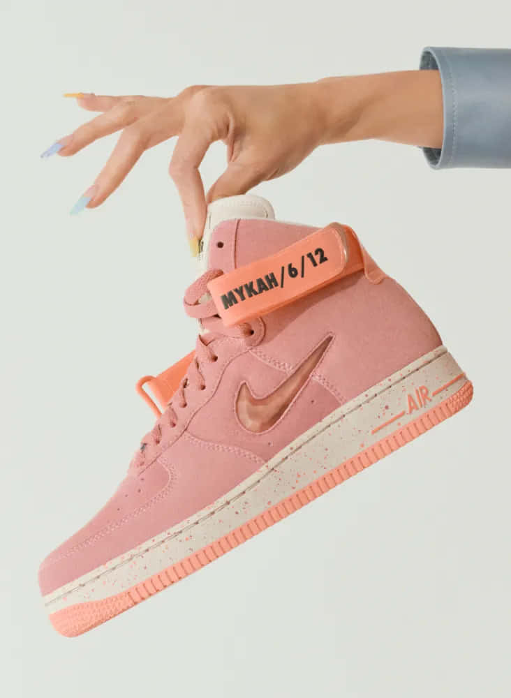 Nike 桌布 壁紙 — 一隻帶有彩色指甲的手，在純色背景上輕輕拿著一雙粉紅色高筒運動鞋。
