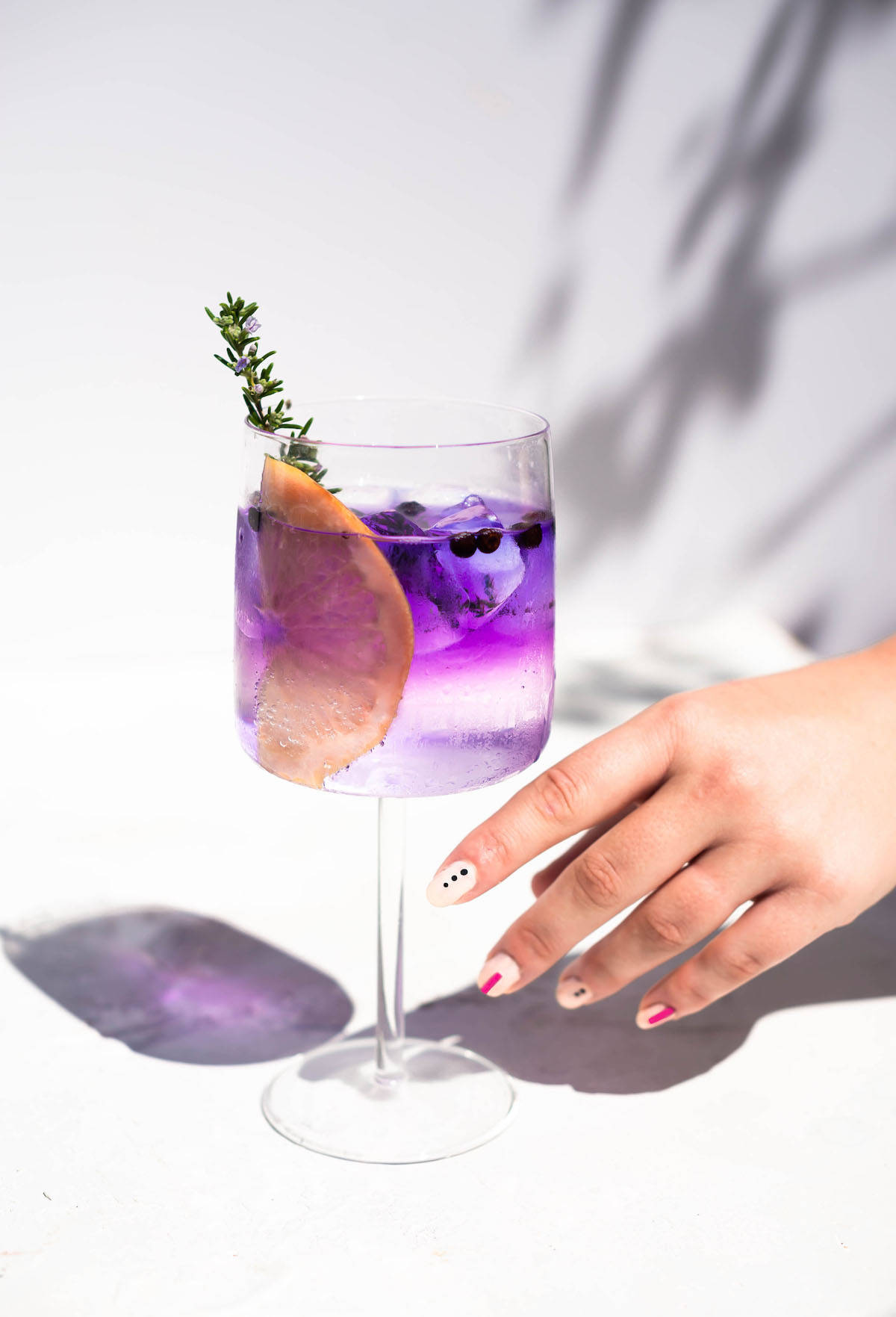 cocktail 桌布 壁紙 — 一隻帶有彩色指甲的手輕輕放在一個充滿活力的紫色雞尾酒旁邊，雞尾酒上裝飾著柑橘和香草，背景是明亮的白色表面。