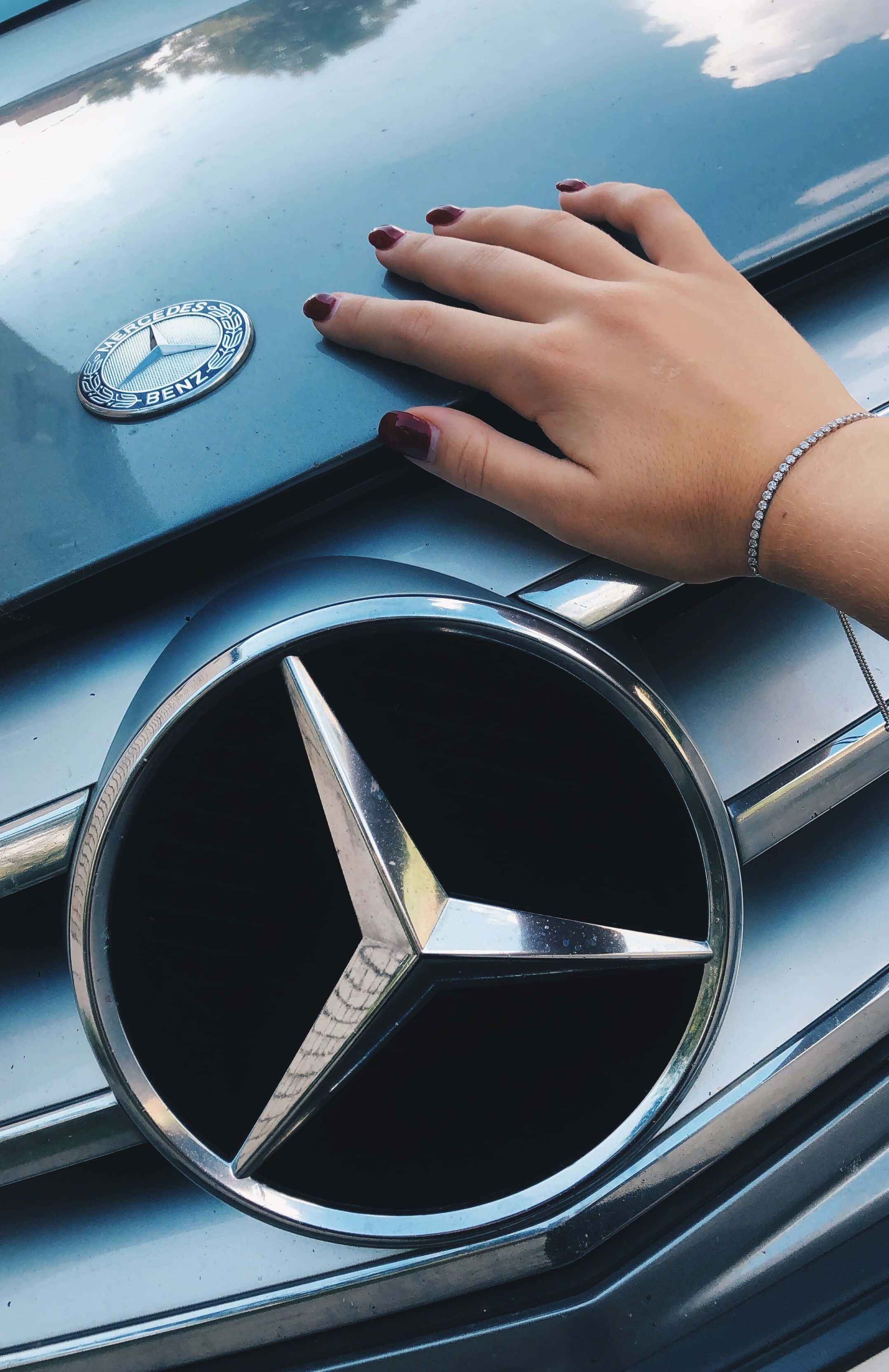 Mercedes 桌布 壁紙 — 一隻帶有深紅色指甲的手輕輕放在一輛時尚的銀色賓士引擎蓋上，突顯標誌性的三叉星徽章。
