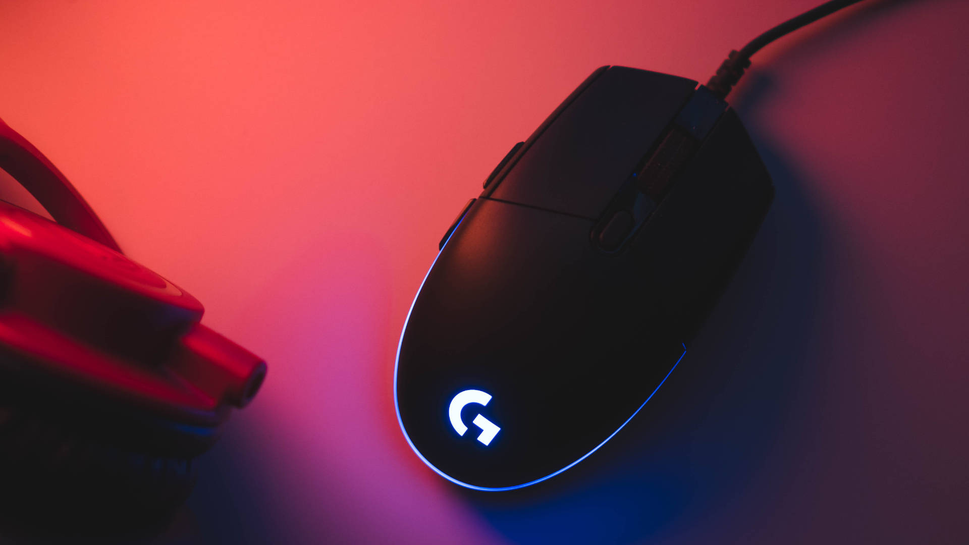 Logitech 桌布 壁紙 — 一隻帶有發光藍色標誌的黑色Logitech遊戲滑鼠，放置在沐浴在紅藍霓虹燈光下的表面上。