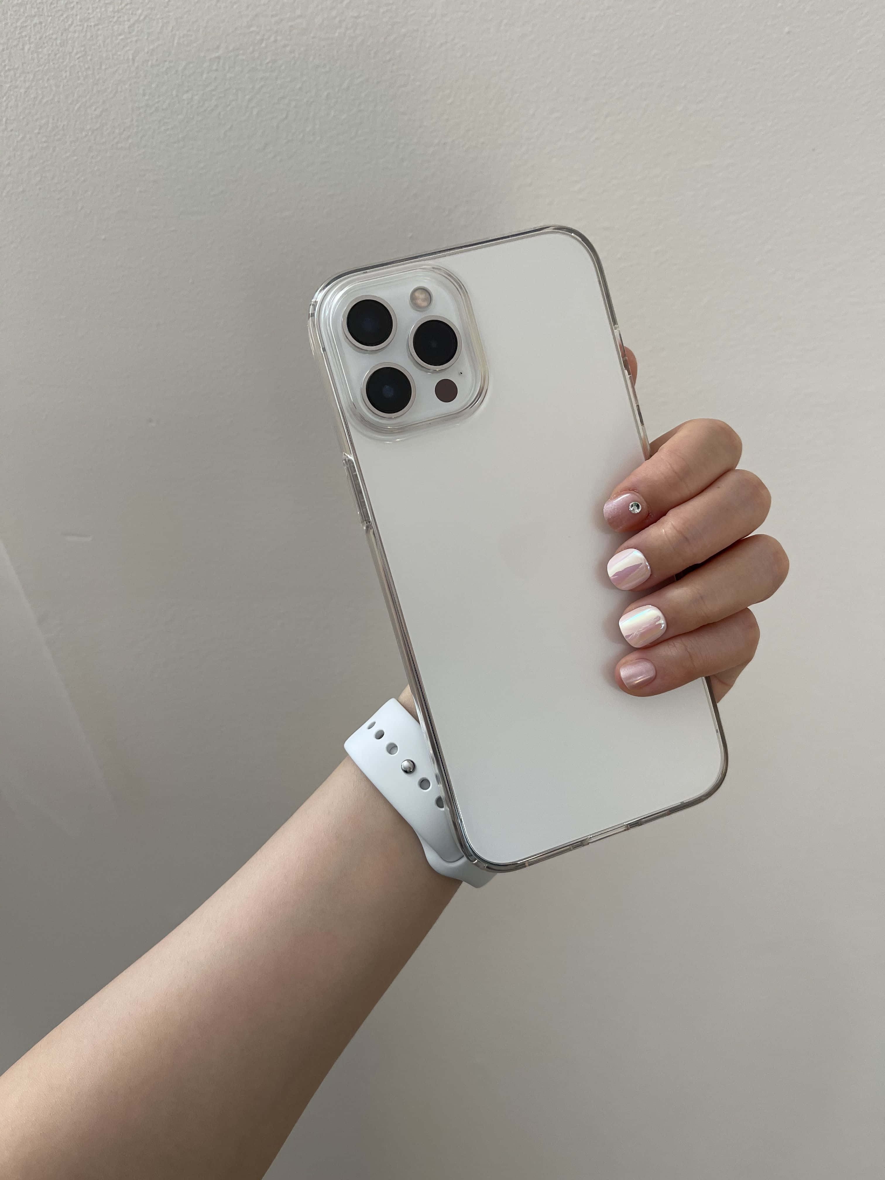 iPhone 12 桌布 壁紙 — 一隻帶有粉紅色亮片指甲的手，拿著一個透明保護殼內的白色iPhone，背景是素色牆壁。