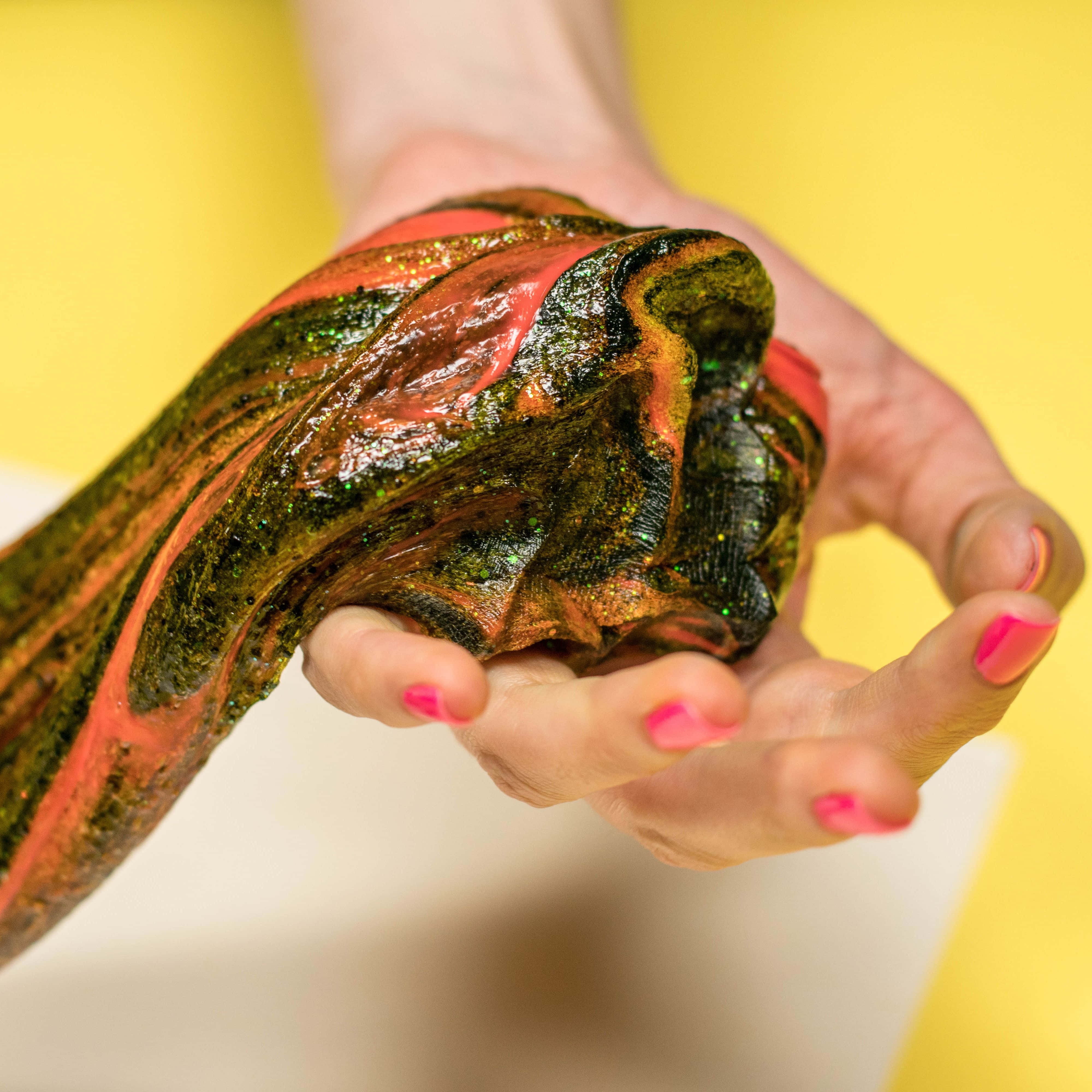 slime 桌布 壁紙 — 一隻帶有粉紅色指甲的手，在明亮的黃色背景前，捧著一個帶有閃亮綠色和橙色漩渦的光澤多色史萊姆。