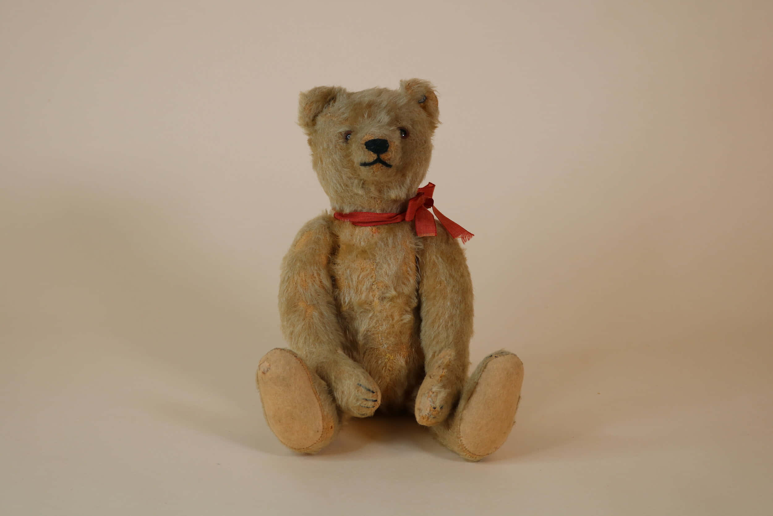 Teddy Bear 桌布 壁紙 — 一隻帶有紅色絲帶的復古磨損泰迪熊，直立在純米色背景前。
