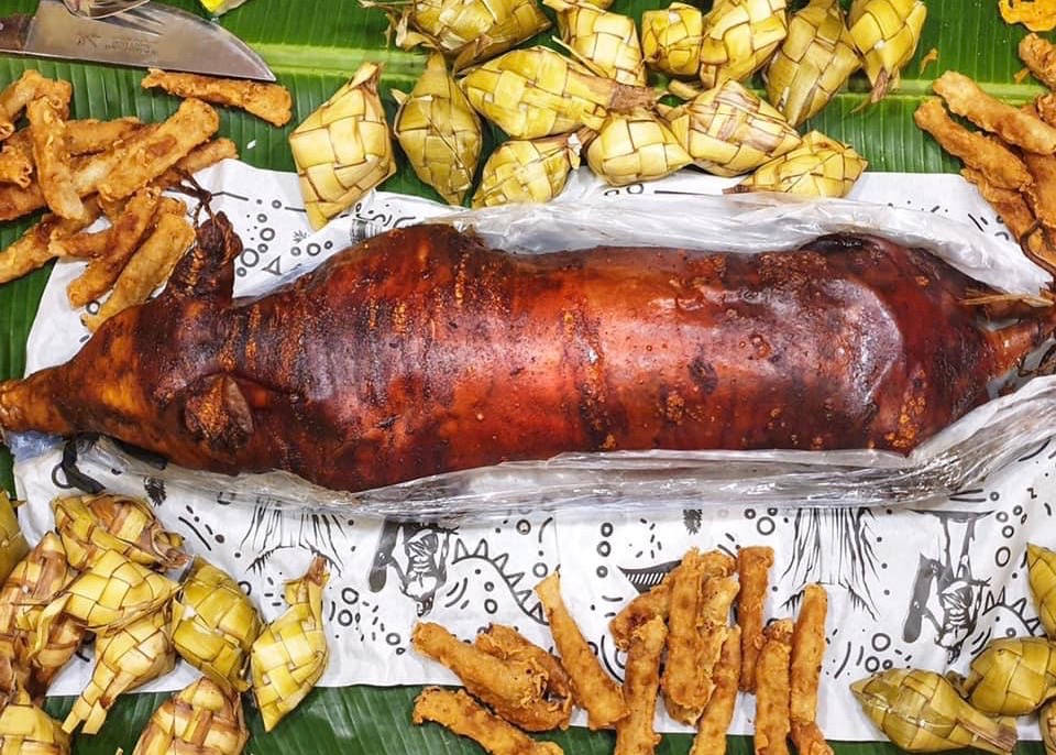 Pig 桌布 壁紙 — 一隻帶有脆皮的整隻烤豬放在香蕉葉上，周圍是編織的香蕉葉包裹和油炸小吃。