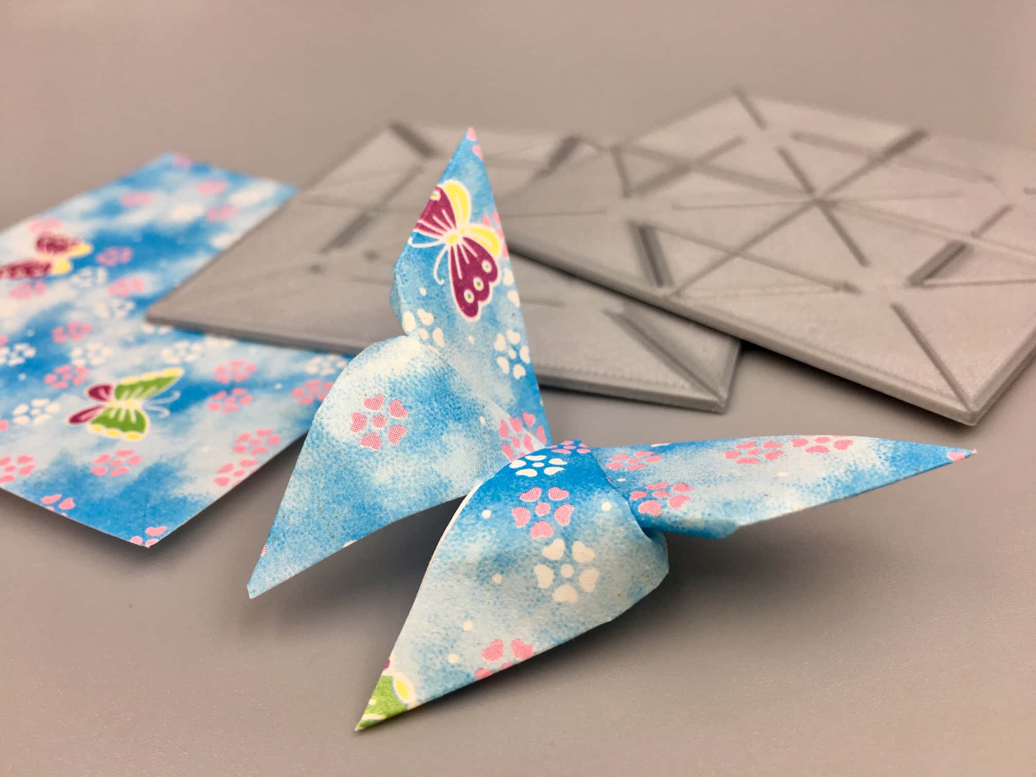 Butterfly Origami 桌布 壁紙 — 一隻帶有花卉圖案的精緻藍色摺紙鳥棲息在折疊紙張和幾何瓷磚旁邊的中性表面上。