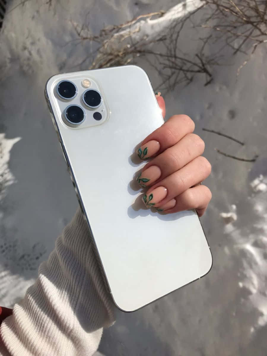 iPhone 12 桌布 壁紙 — 一隻帶有葉子指甲藝術的手，在雪景背景中拿著一部白色智慧型手機，背景有光禿的樹枝。