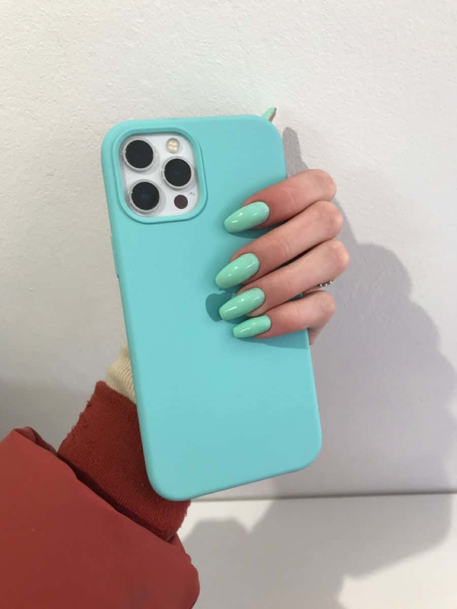 iPhone 12 桌布 壁紙 — 一隻帶有薄荷綠色指甲的手拿著淺藍色手機殼，靠在純白牆上。