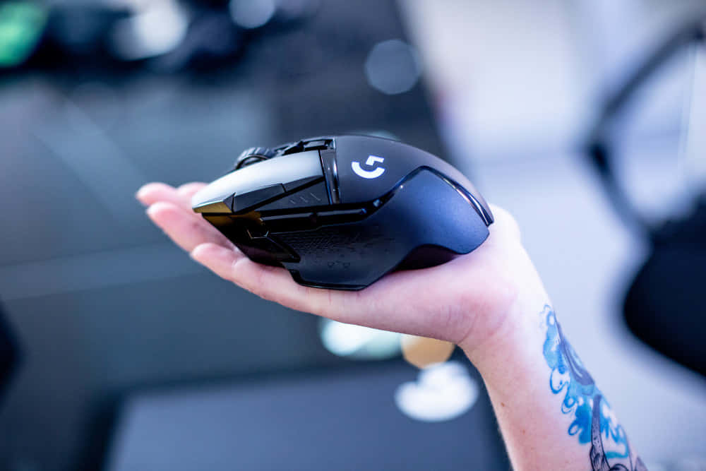 Gaming Mouse 桌布 壁紙 — 一隻帶有藍色刺青的手，在模糊的科技工作空間背景下，握著一款時尚的黑色Logitech遊戲滑鼠。