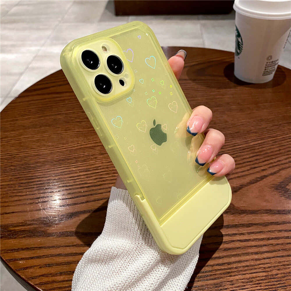 iPhone 美學 桌布 壁紙 — 一隻帶有藍色指尖指甲的手，在木桌上拿著一個帶有心形貼紙的黃色手機殼，旁邊放著一杯咖啡。