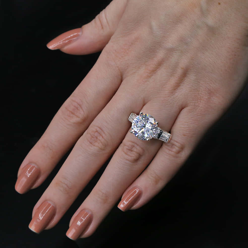 rings 桌布 壁紙 — 一隻帶有裸色指甲的手在黑色背景上展示一枚閃閃發光的單鑽訂婚戒指。