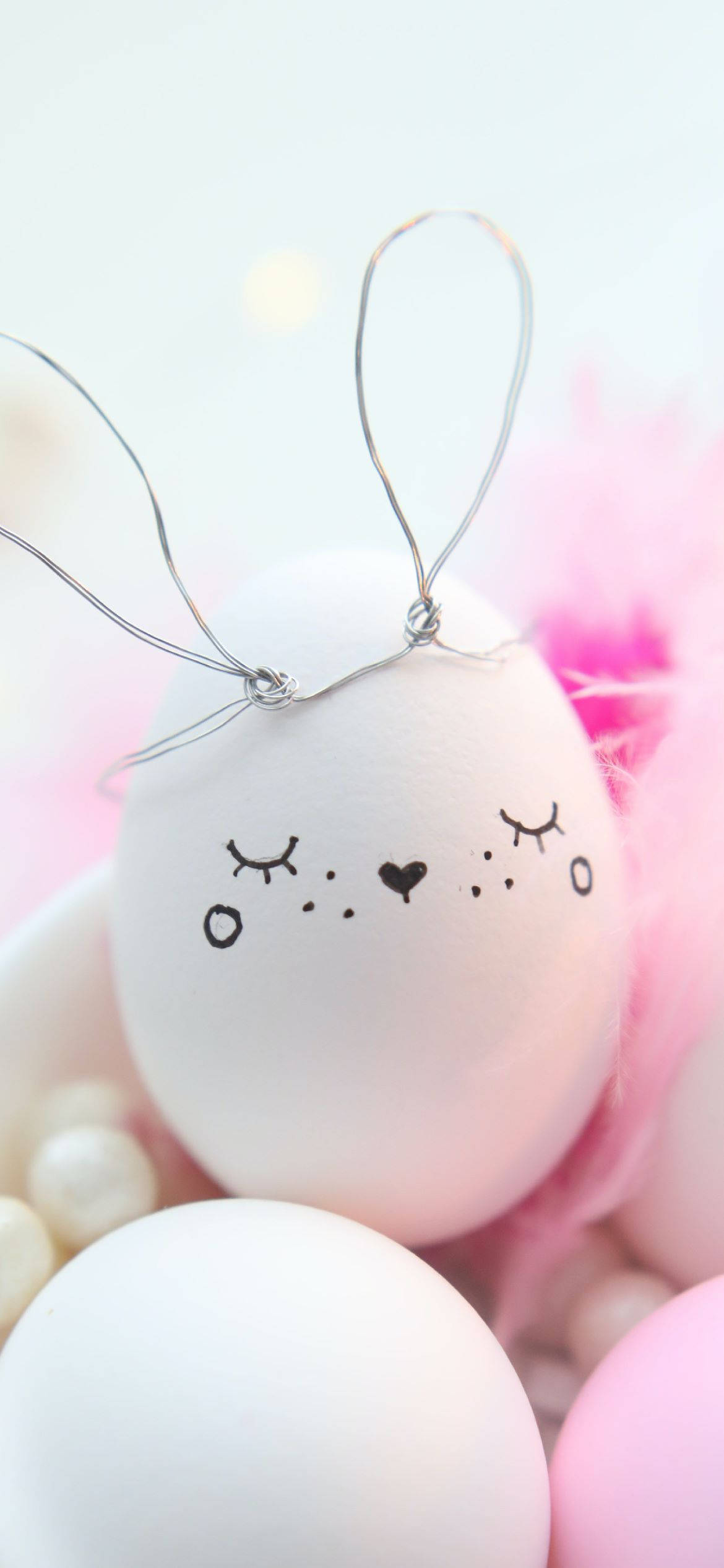 Easter Phone 桌布 壁紙 — 一隻帶有鐵絲兔耳和可愛黑色墨水臉的白蛋，放置在粉紅色絨毛物料和珍珠之間。