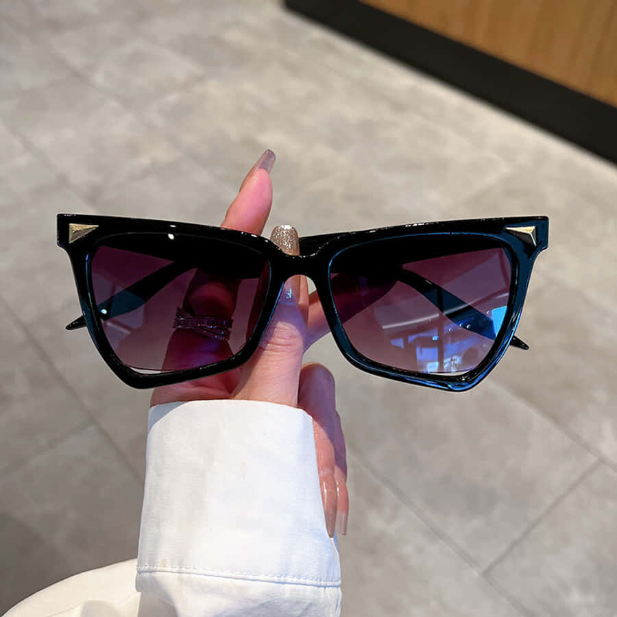 Purple Sunglasses 桌布 壁紙 — 一隻帶有閃亮指甲的手，在瓷磚地板背景下，拿著一副大膽的黑色貓眼太陽眼鏡，配紫色鏡片。